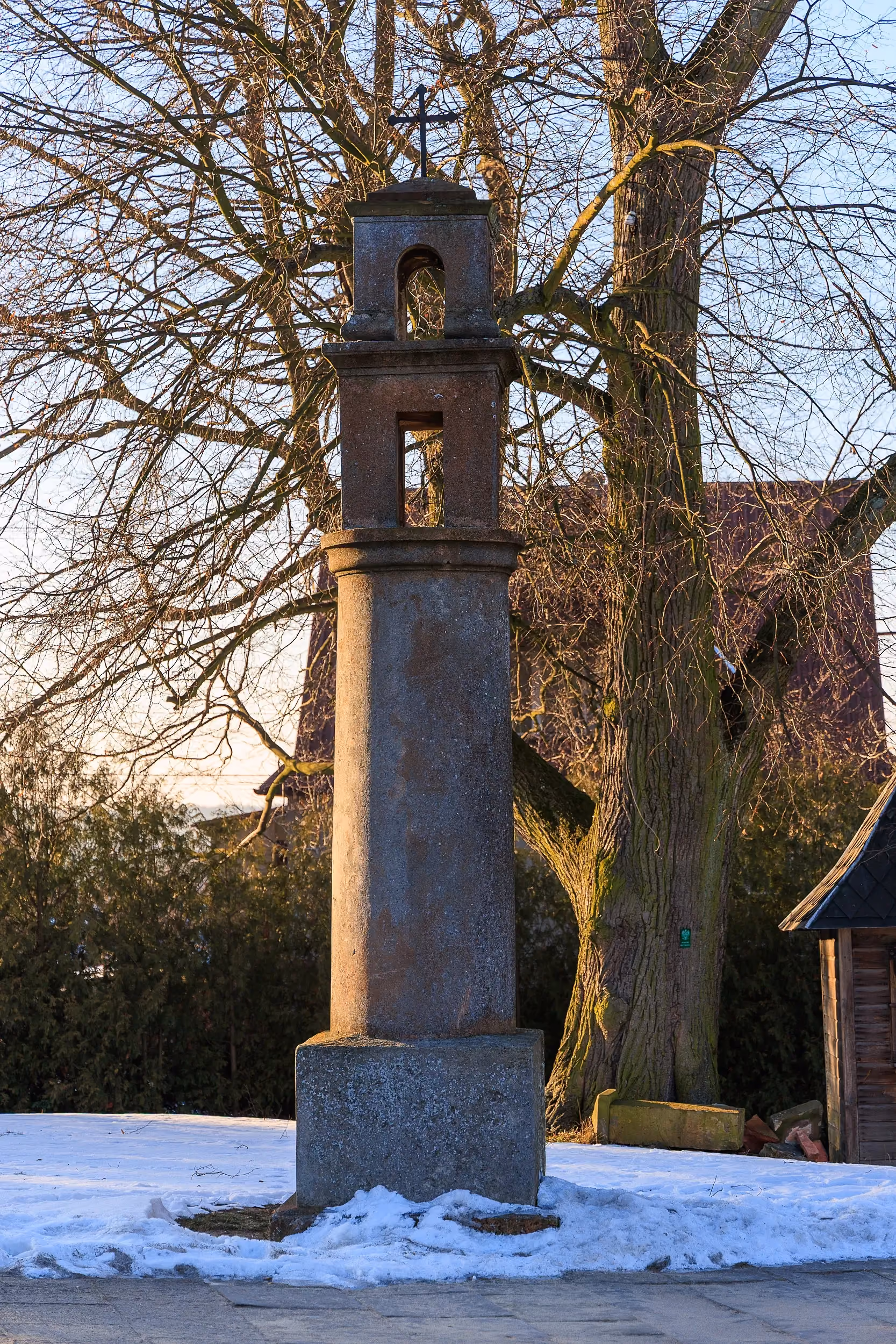 Kamienna kapliczka słupowa przy kościele w Przedborzu. Smukły monument z wnękami i krzyżem na szczycie, otoczony drzewami oraz zimowym krajobrazem.