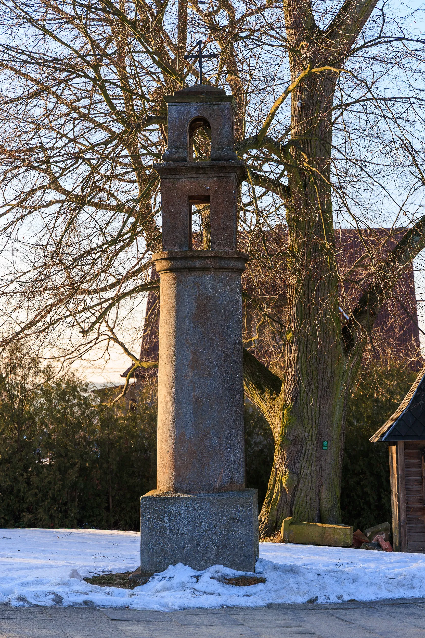 Kamienna kapliczka słupowa przy kościele w Przedborzu. Smukły monument z wnękami i krzyżem na szczycie, otoczony drzewami oraz zimowym krajobrazem.
