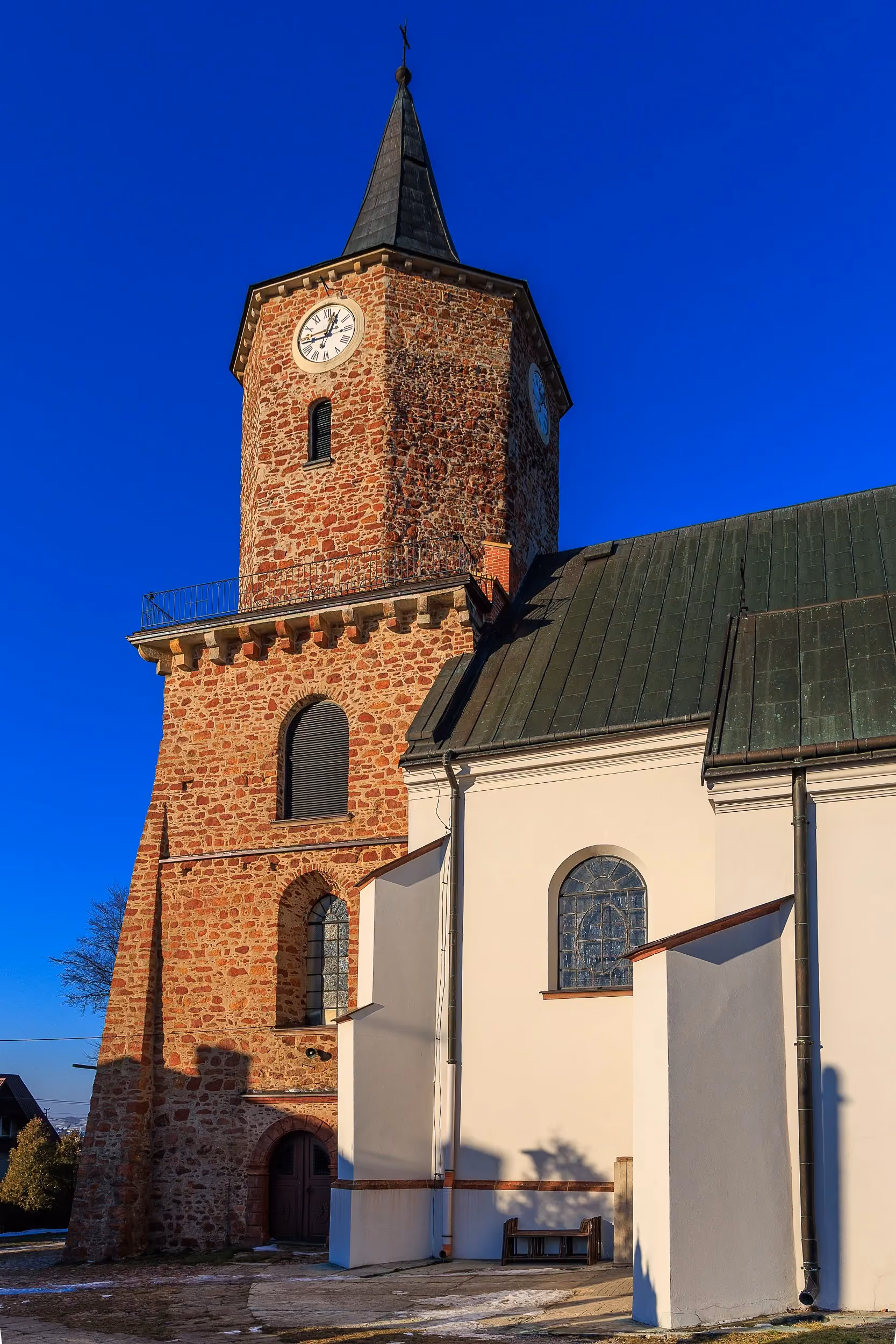 Kościół św. Aleksego w Przedborzu. Widok na ceglaną wieżę zegarową oraz białą nawę świątyni z wysokimi oknami i metalowym dachem.