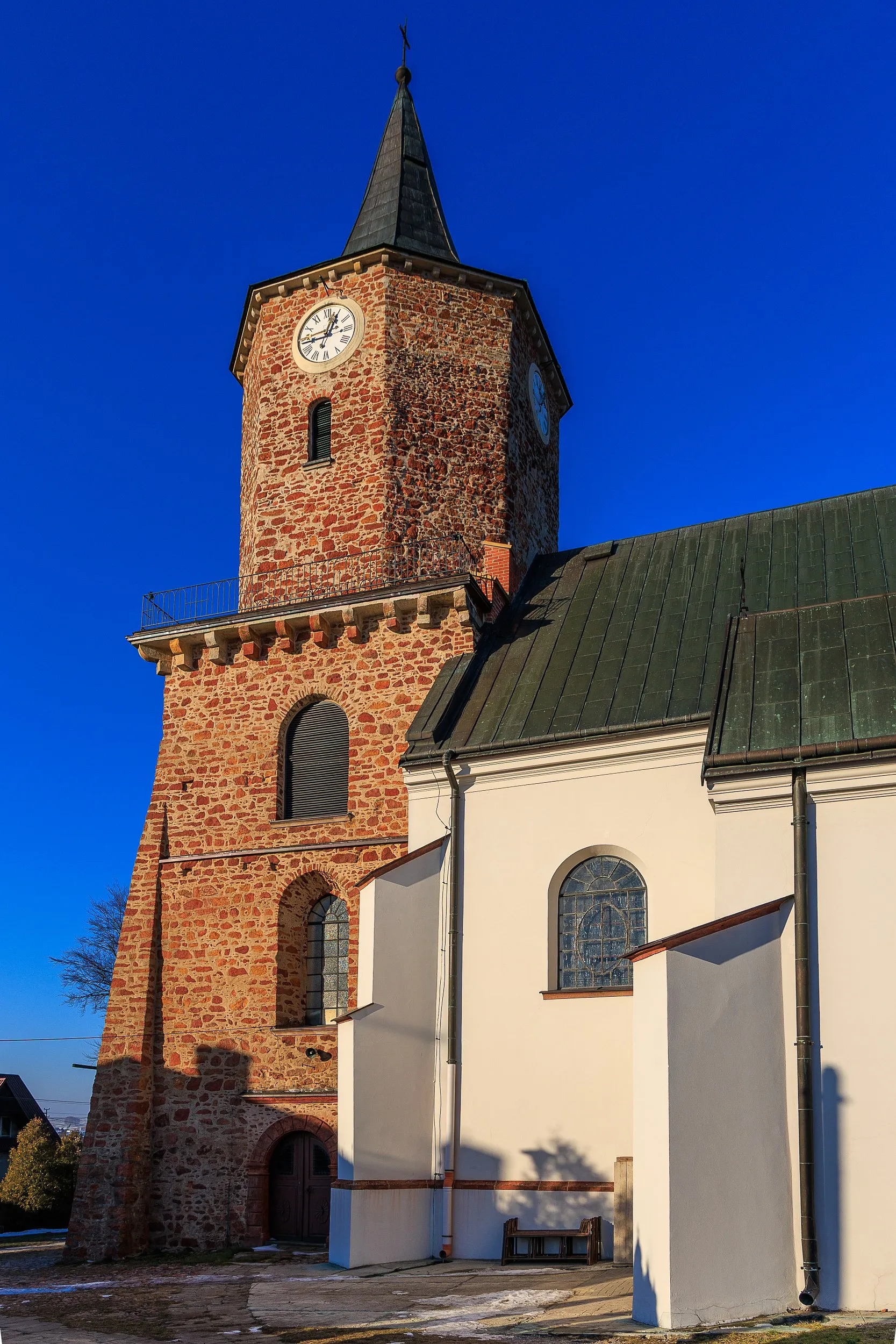 Kościół św. Aleksego w Przedborzu. Widok na ceglaną wieżę zegarową oraz białą nawę świątyni z wysokimi oknami i metalowym dachem.
