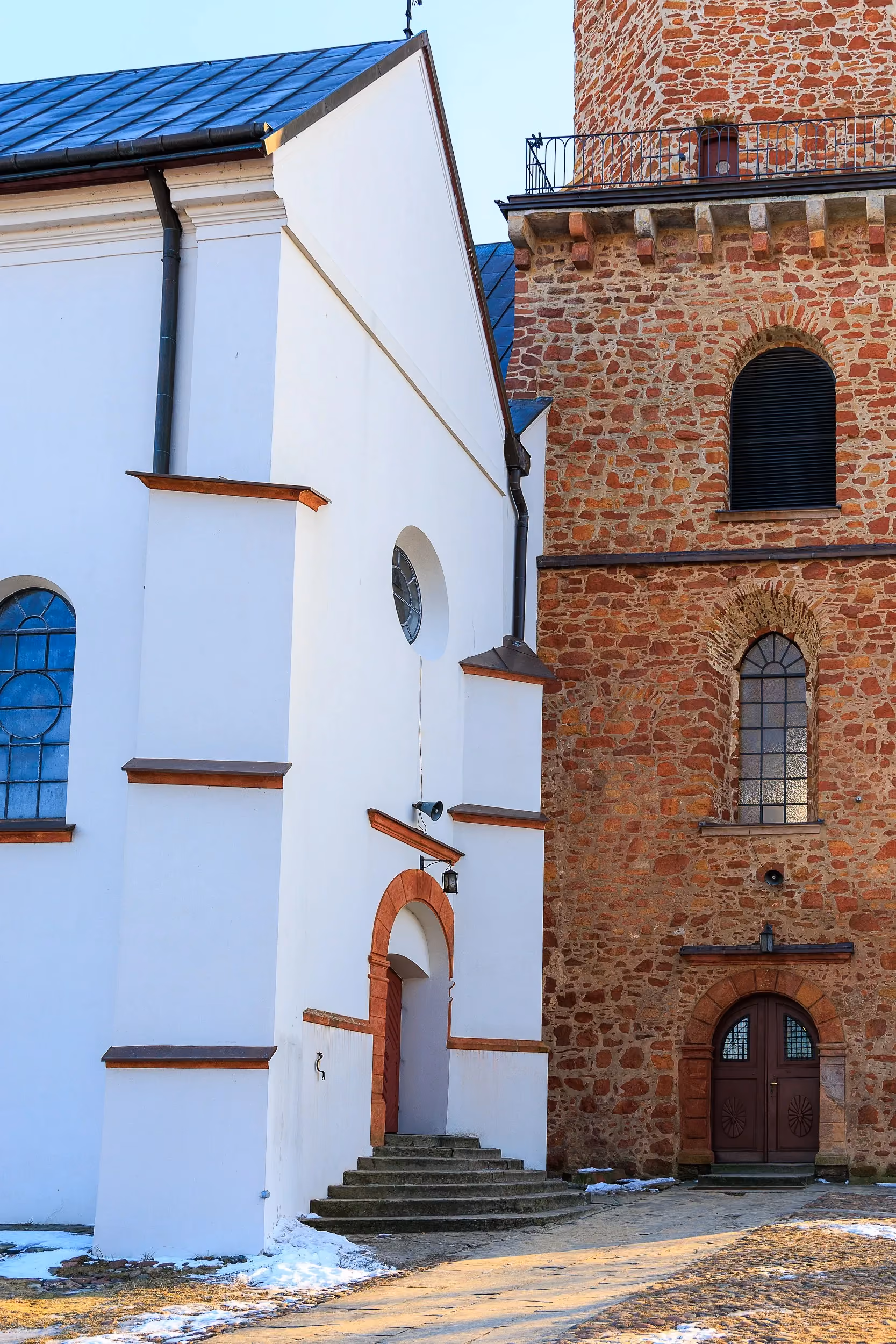 Kościół św. Aleksego w Przedborzu. Widok na ceglaną wieżę zegarową oraz białą nawę świątyni z wysokimi oknami i metalowym dachem.