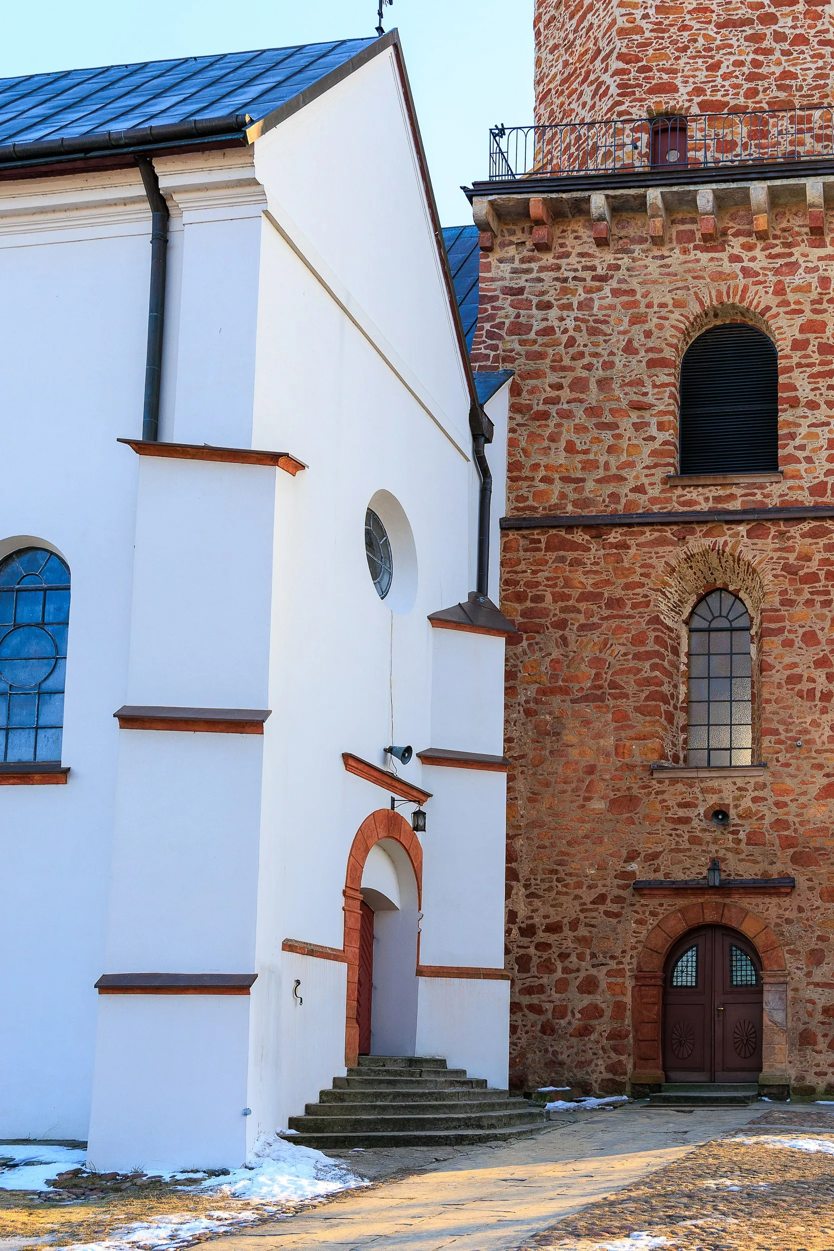 Kościół św. Aleksego w Przedborzu. Widok na ceglaną wieżę zegarową oraz białą nawę świątyni z wysokimi oknami i metalowym dachem.