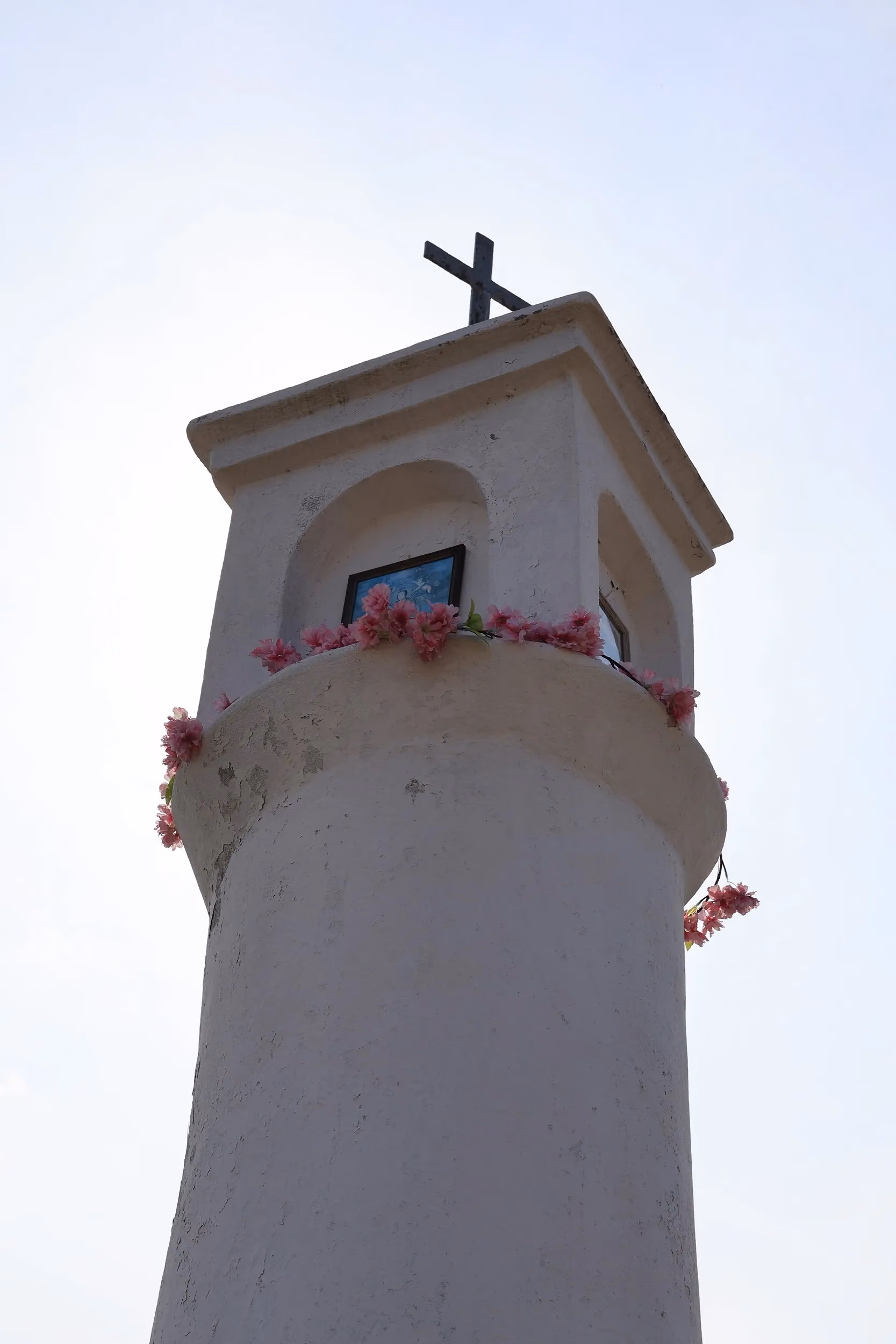 Detal górnej części kapliczki słupowej. Widoczne wnęki z obrazkami religijnymi, ozdobne kwiaty oraz krzyż wieńczący konstrukcję.