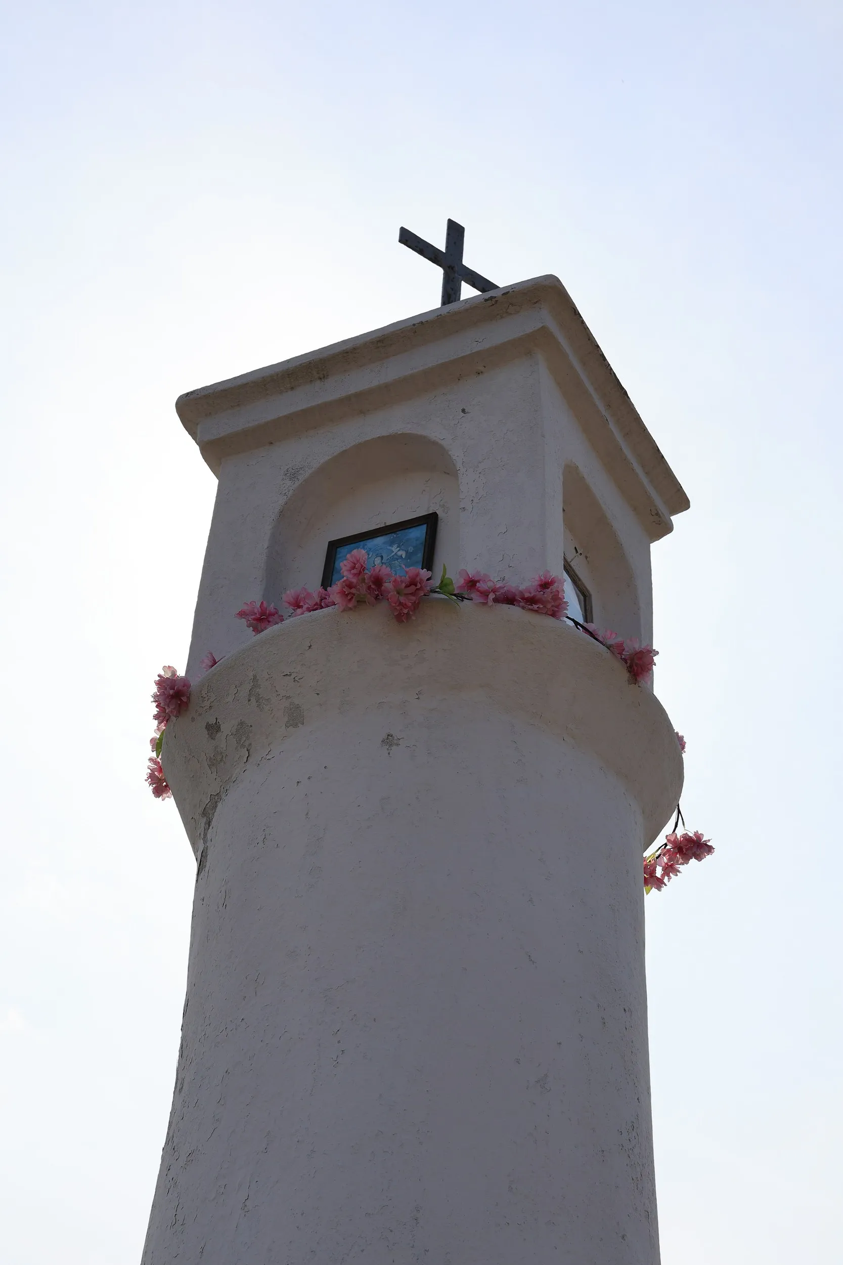 Detal górnej części kapliczki słupowej. Widoczne wnęki z obrazkami religijnymi, ozdobne kwiaty oraz krzyż wieńczący konstrukcję.