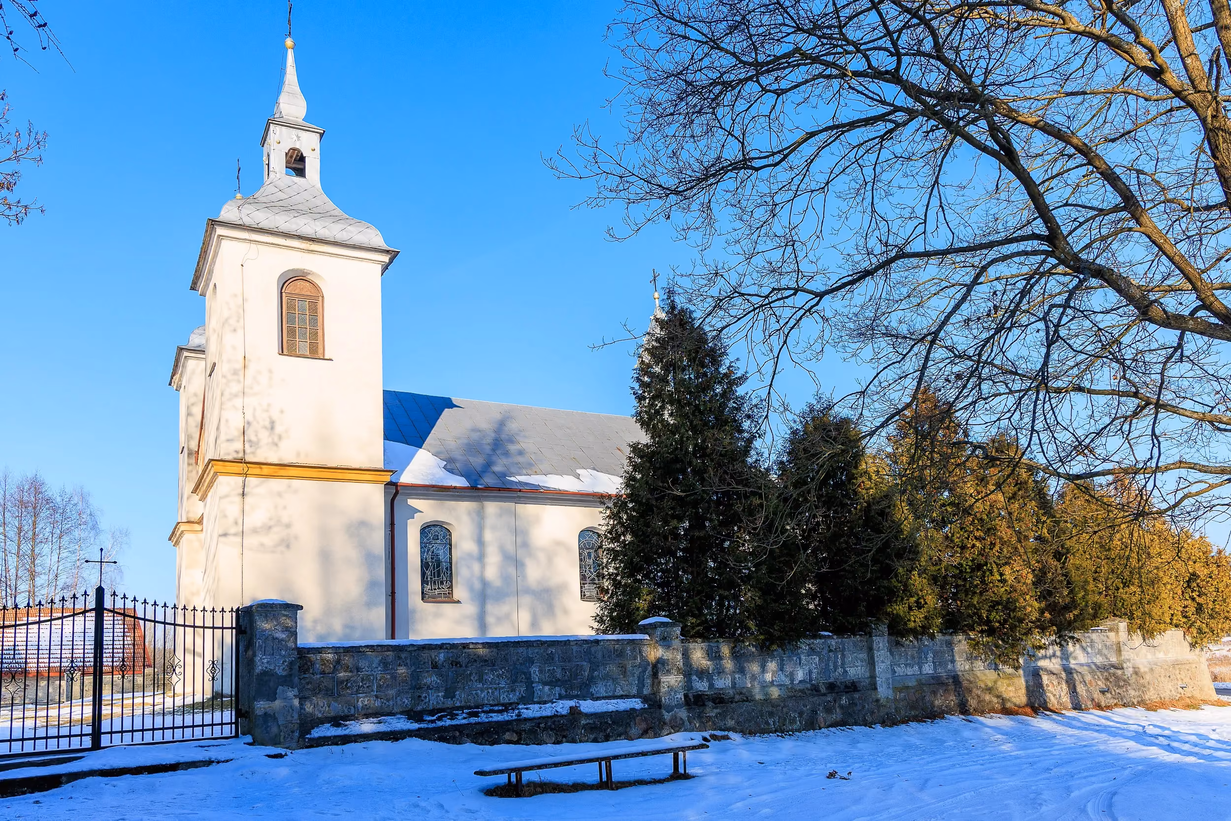 Kościół w Żeleźnicy widziany od strony ogrodzenia. Jasna świątynia z wieżą i dachem pokrytym śniegiem na tle bezchmurnego nieba.
