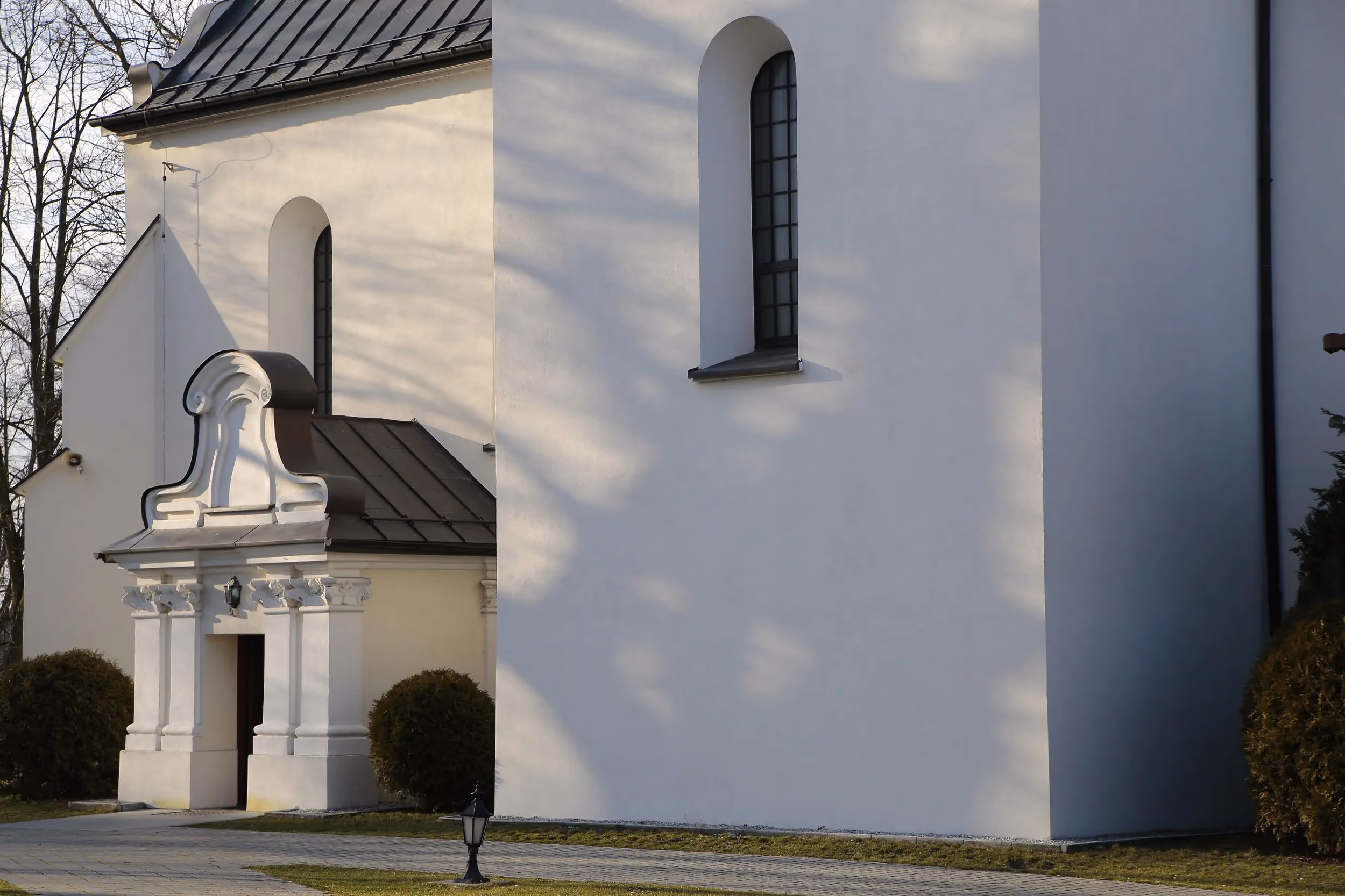 Fragment kościoła w Strzałkowie z bocznym wejściem. Widoczne wysokie okna, jasna elewacja oraz ozdobny portal wejściowy.