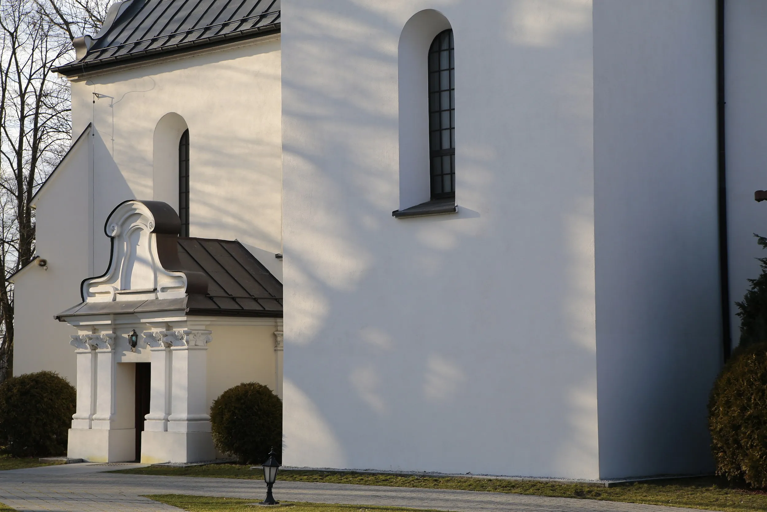 Fragment kościoła w Strzałkowie z bocznym wejściem. Widoczne wysokie okna, jasna elewacja oraz ozdobny portal wejściowy.