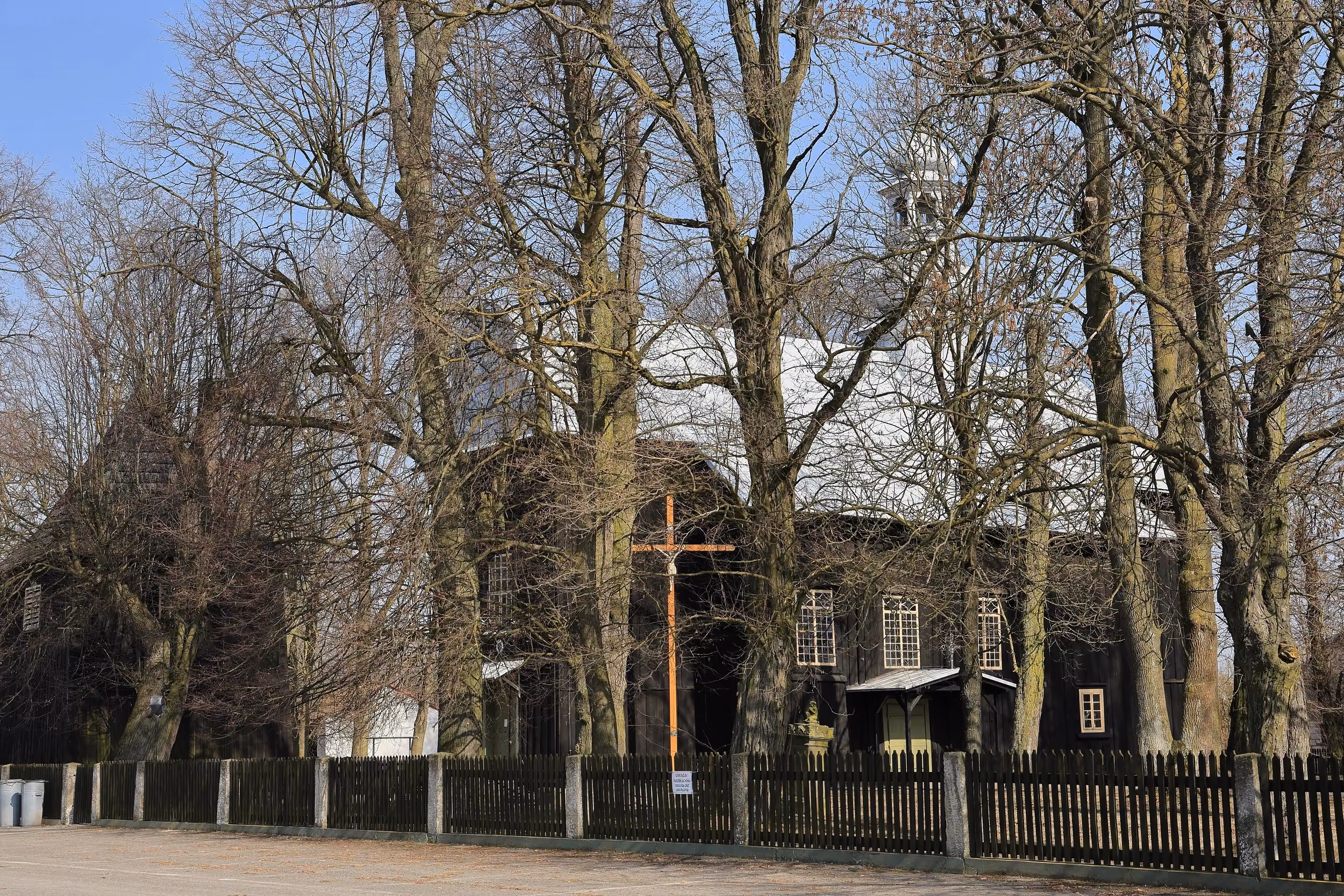 Drewniany kościół w Niedośpielinie widziany zza ogrodzenia. Budynek otaczają wysokie drzewa i przykościelny teren.