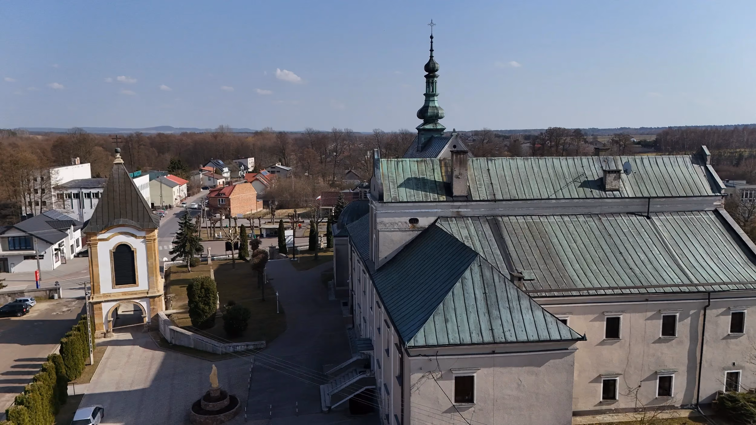 Widok z lotu ptaka na kościół i dzwonnicę w Wielgomłynach. Widoczne są dachy budynków, dziedziniec oraz okolica miejscowości.