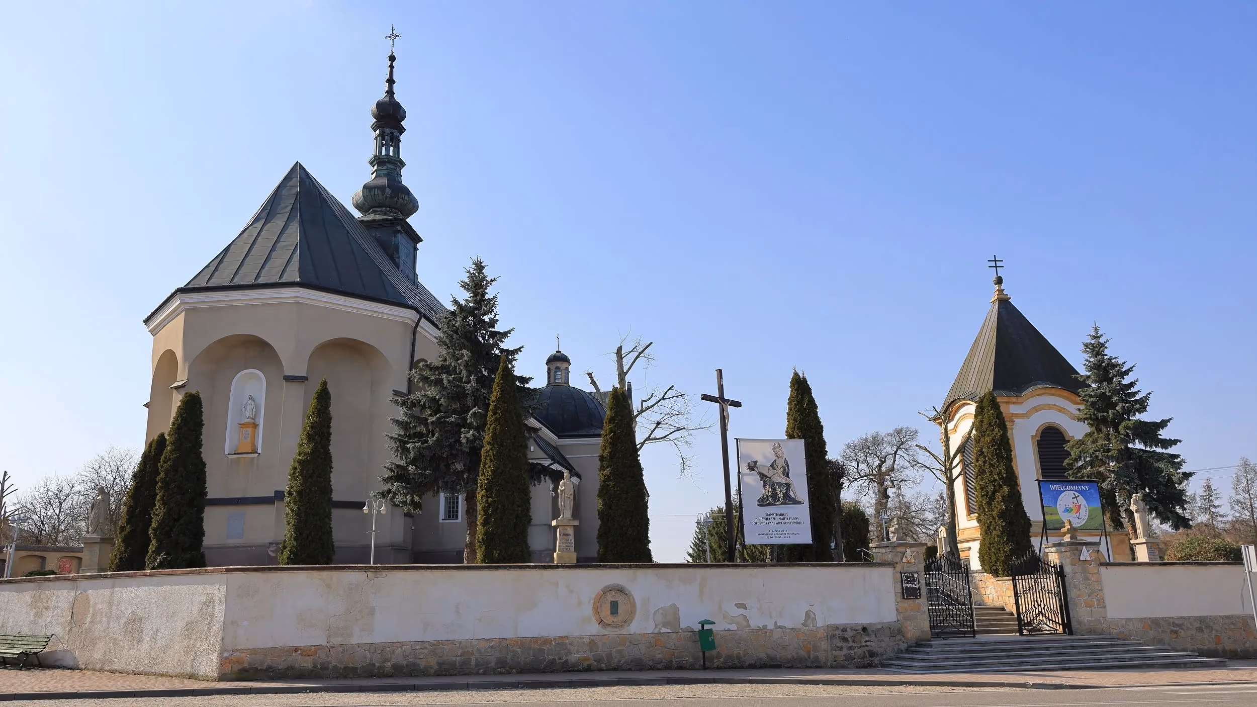 Widok kościoła i dzwonnicy w Wielgomłynach od strony ulicy. Kompleks otacza mur, ozdobna brama i szpaler drzew.