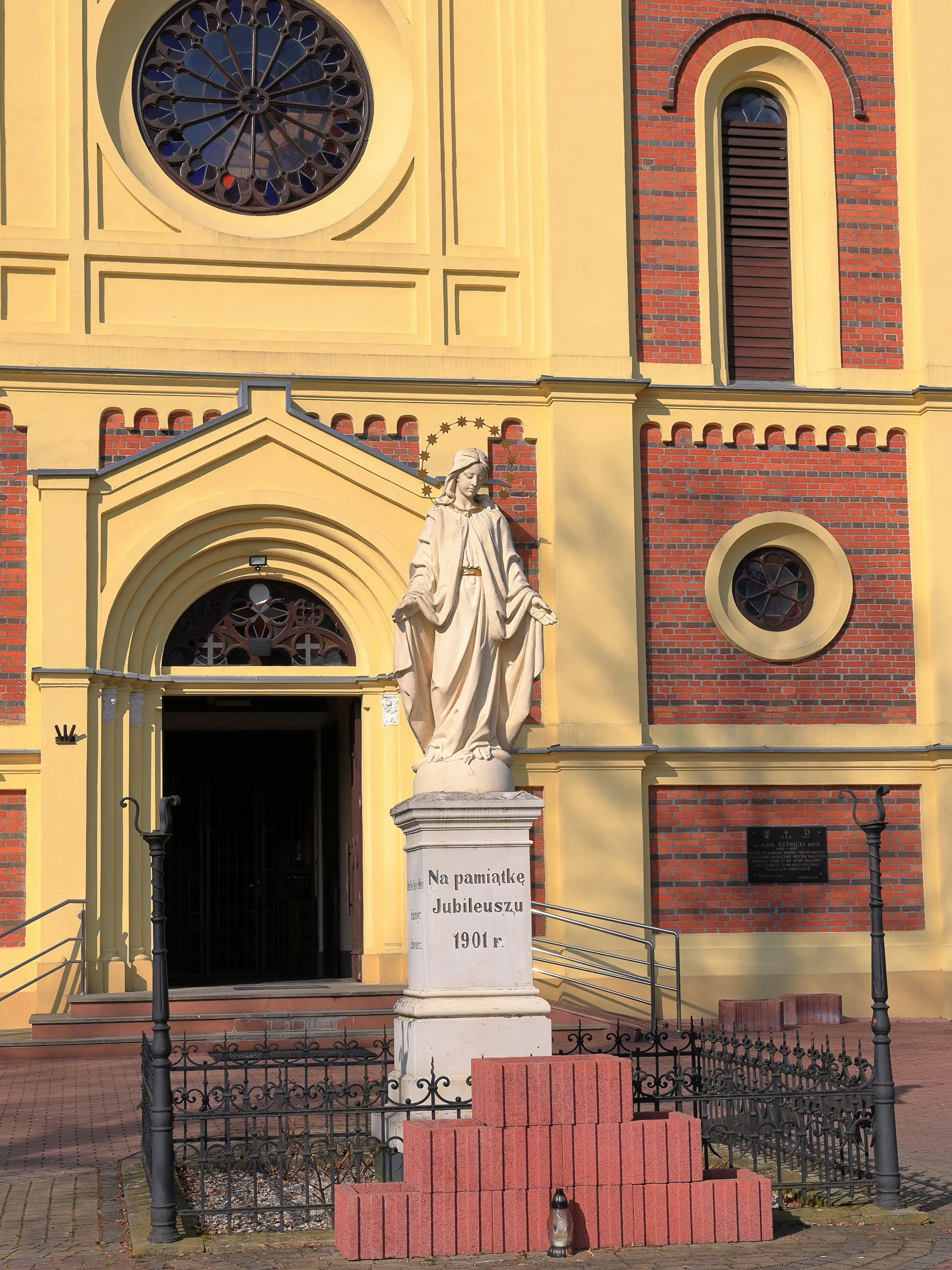 Figura Matki Bożej przed kościołem w Żytnie. Pomnik stoi przed głównym wejściem do świątyni o ceglanej i jasnej elewacji.