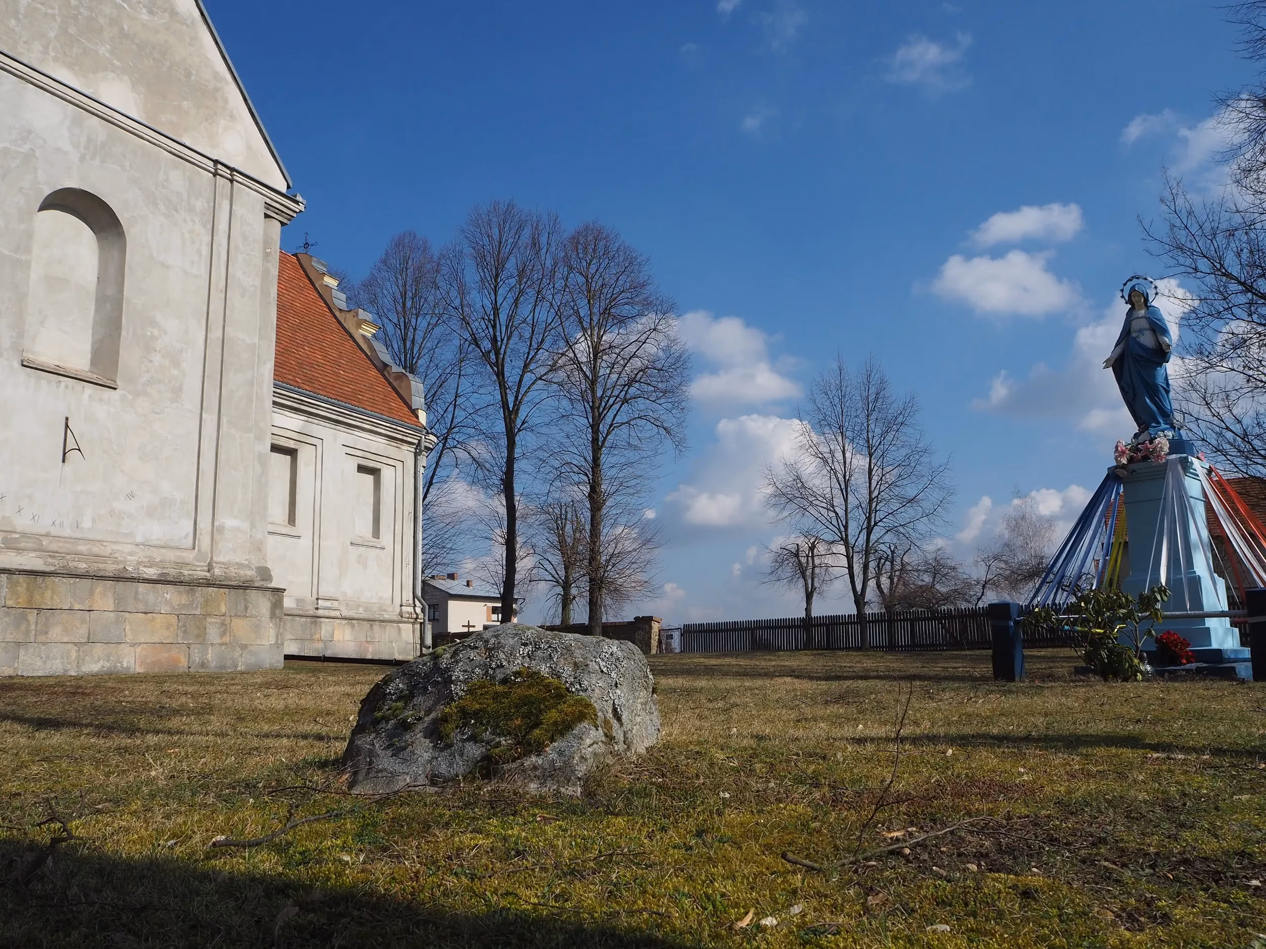 Boczny widok kościoła w Maluszynie. Na trawniku przed świątynią widoczny głaz narzutowy oraz figura Matki Bożej na postumencie.