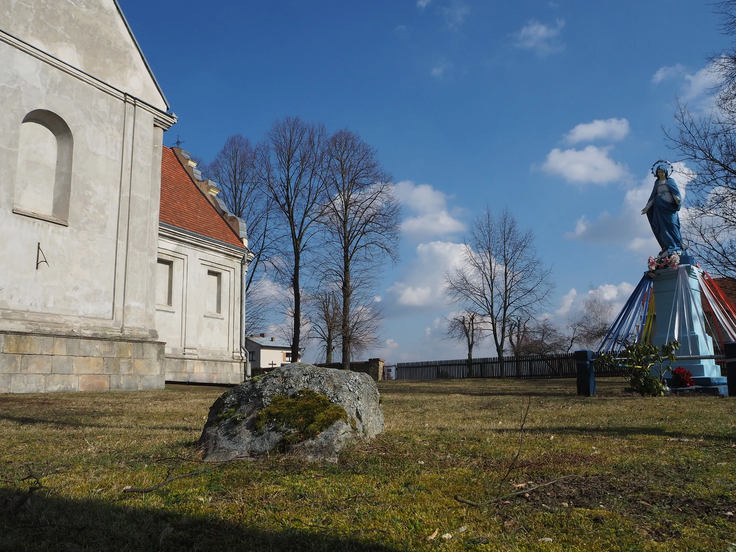 Boczny widok kościoła w Maluszynie. Na trawniku przed świątynią widoczny głaz narzutowy oraz figura Matki Bożej na postumencie.