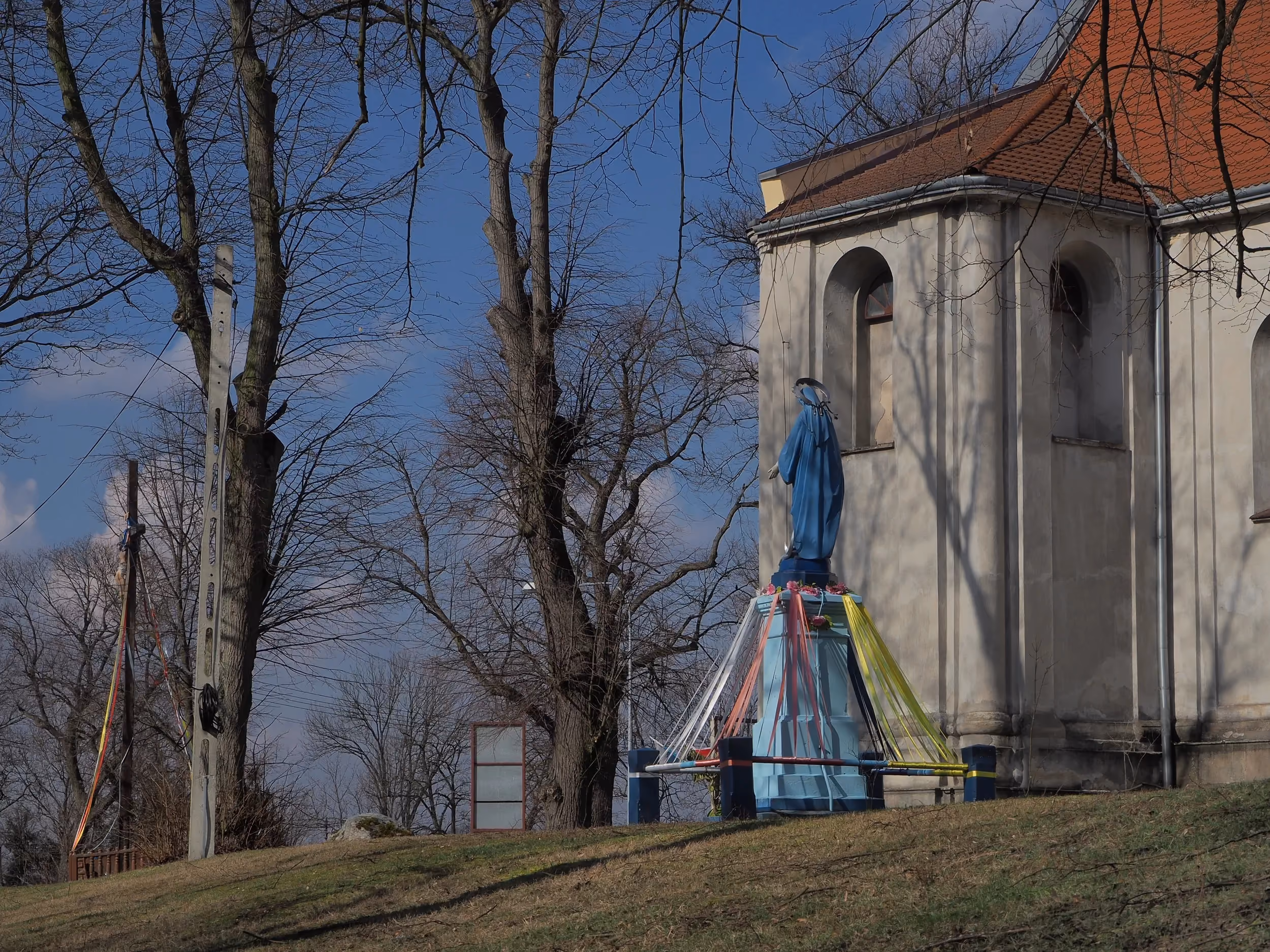Figura Matki Bożej ustawiona obok kościoła w Maluszynie. W tle widoczne drzewa, krzyż oraz fragment świątyni.