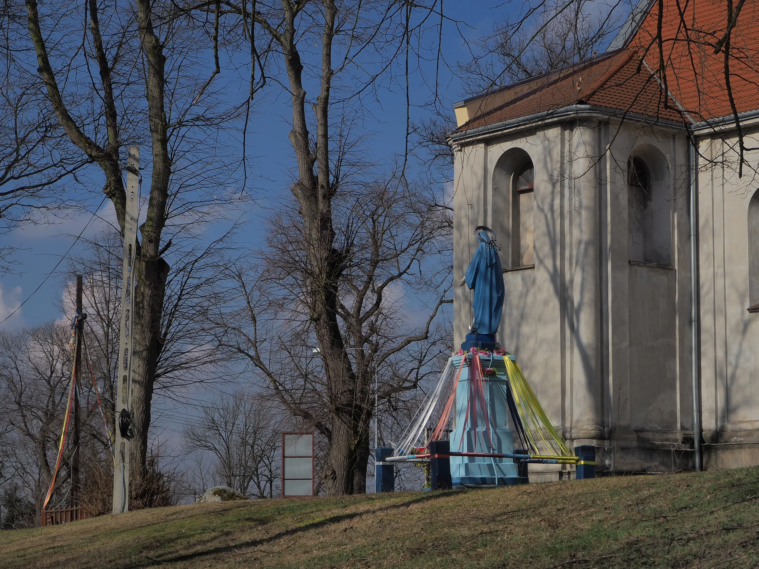 Figura Matki Bożej ustawiona obok kościoła w Maluszynie. W tle widoczne drzewa, krzyż oraz fragment świątyni.