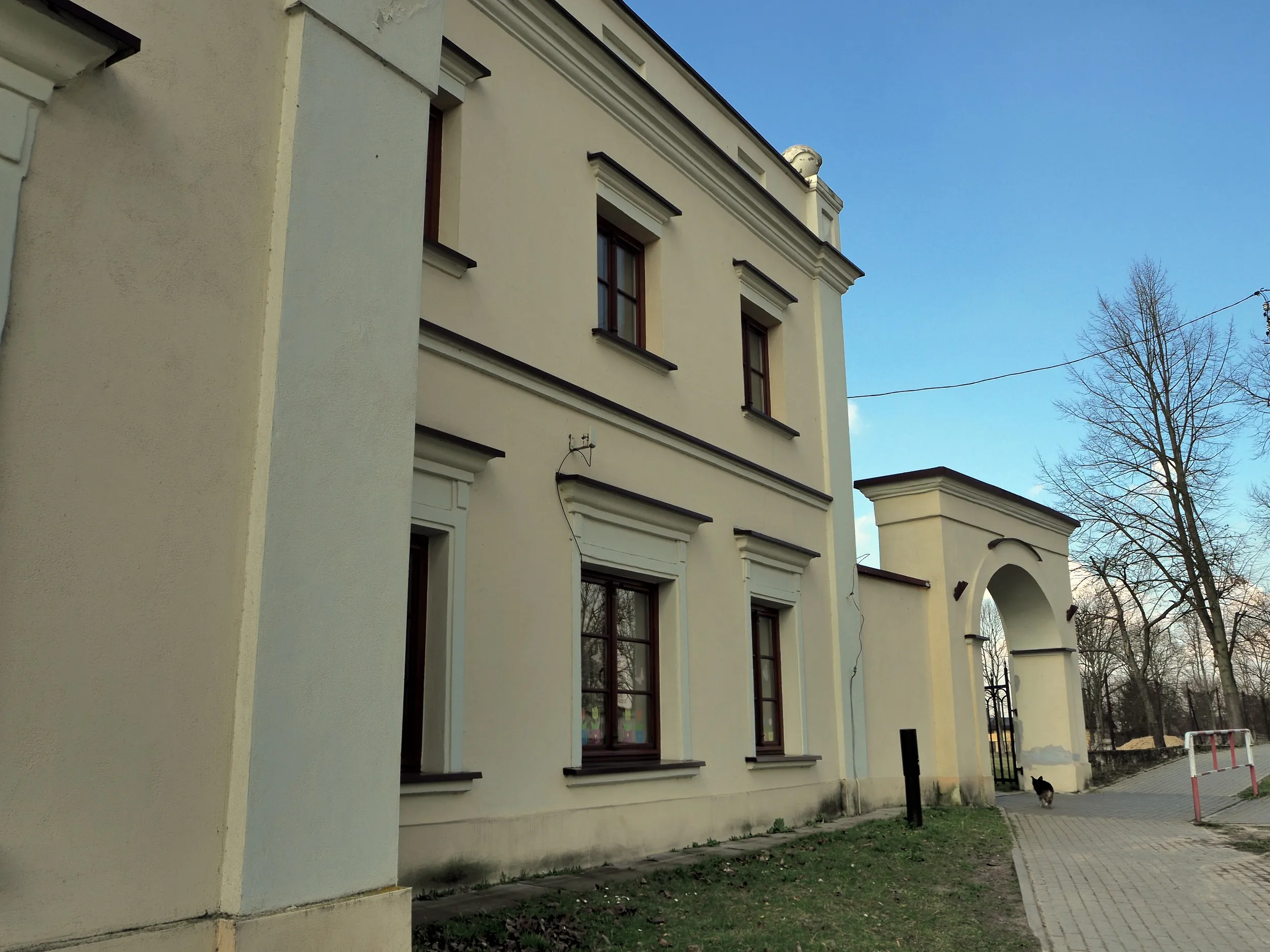 Boczna elewacja historycznego budynku w Maluszynie. W tle widoczna brama wjazdowa prowadząca na teren dawnego zespołu pałacowego.