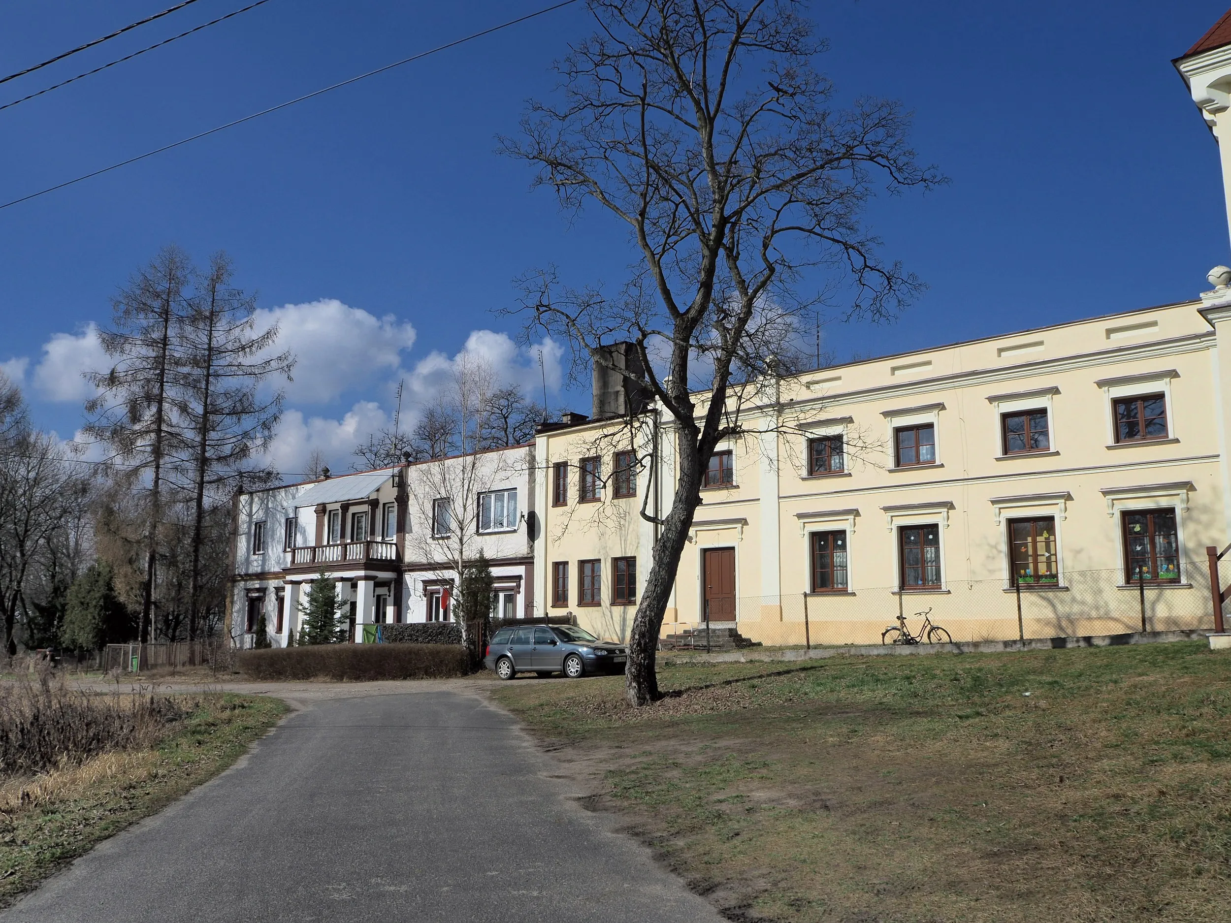 Zabudowania zespołu pałacowego w Maluszynie. Przy drodze widoczne są historyczne budynki otoczone drzewami oraz niewielki plac przed obiektem.
