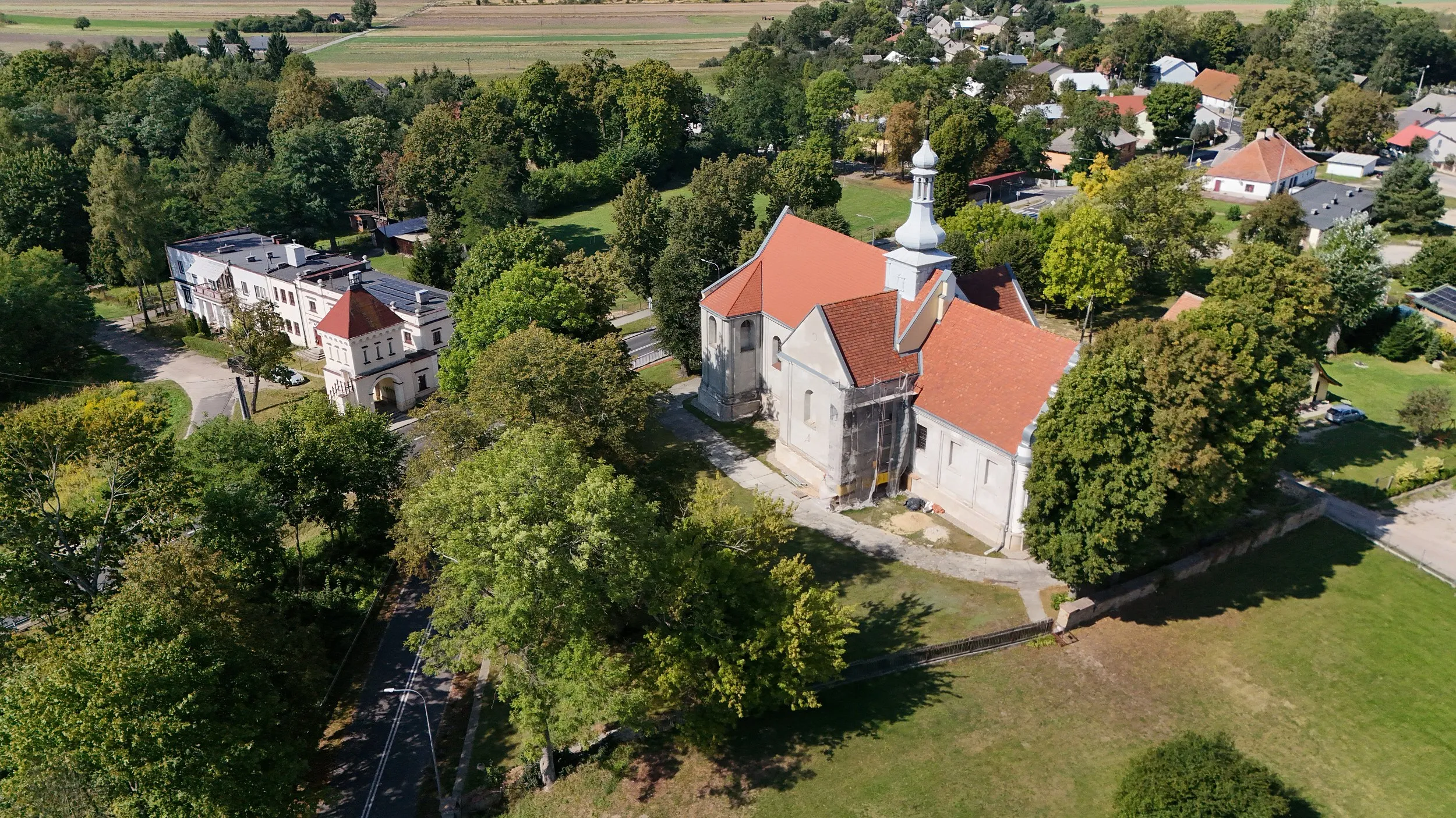 Widok z lotu ptaka na kościół i zabudowania w Maluszynie. Świątynia z czerwonym dachem otoczona jest drzewami, a obok widoczne są historyczne budynki dawnego zespołu dworskiego.