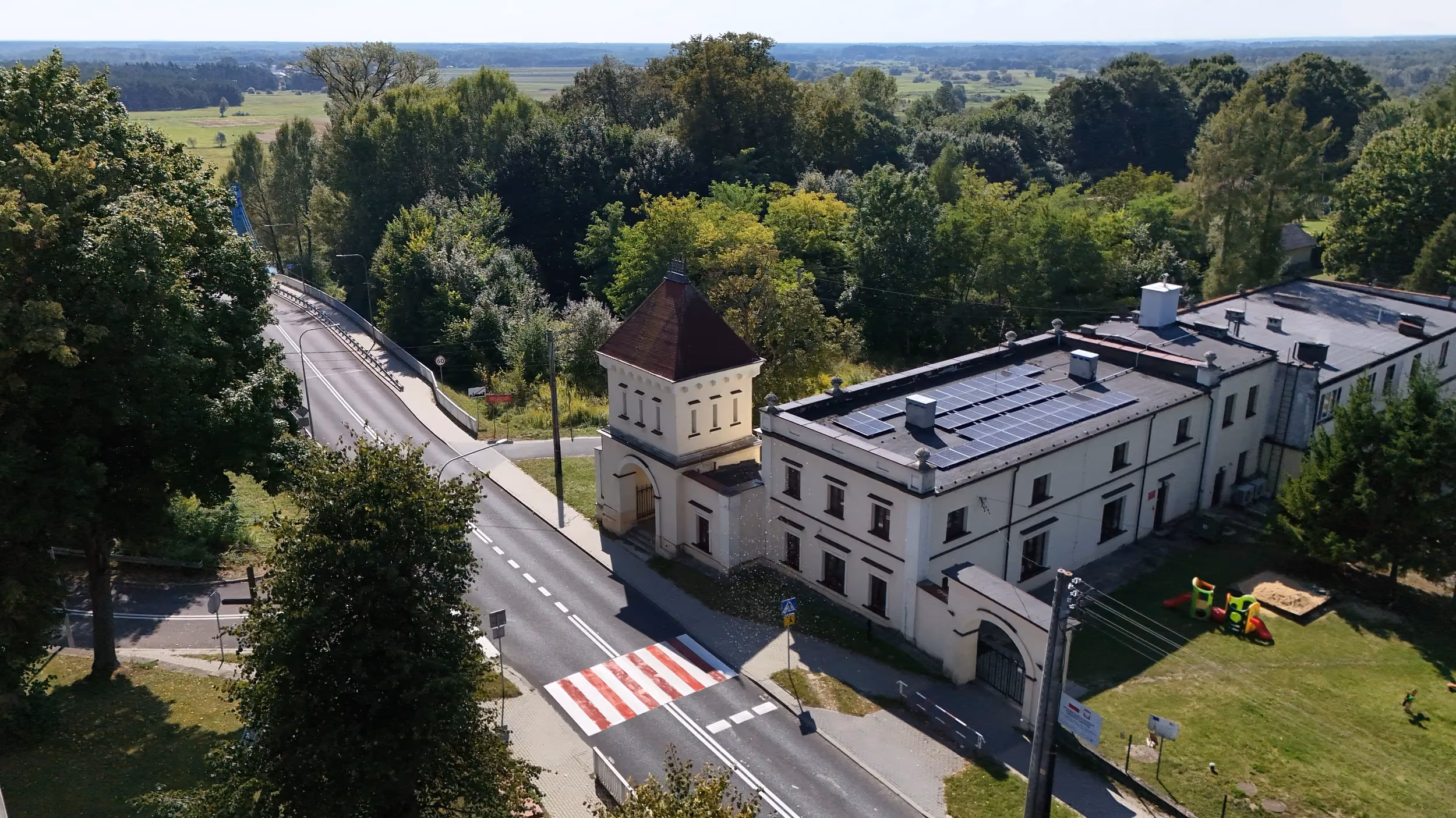 Widok z lotu ptaka na zabytkowy zespół pałacowy w Maluszynie. Widoczna jest wieża bramna, historyczny budynek oraz otaczające je drzewa, droga i most nad rzeką.