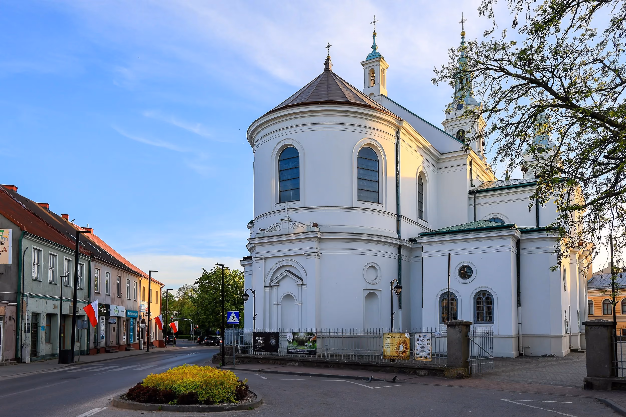 Widok bocznej elewacji Kolegiaty św. Lamberta w Radomsku. Biała świątynia z półkolistą apsydą i wieżami góruje nad zabudową centrum miasta.