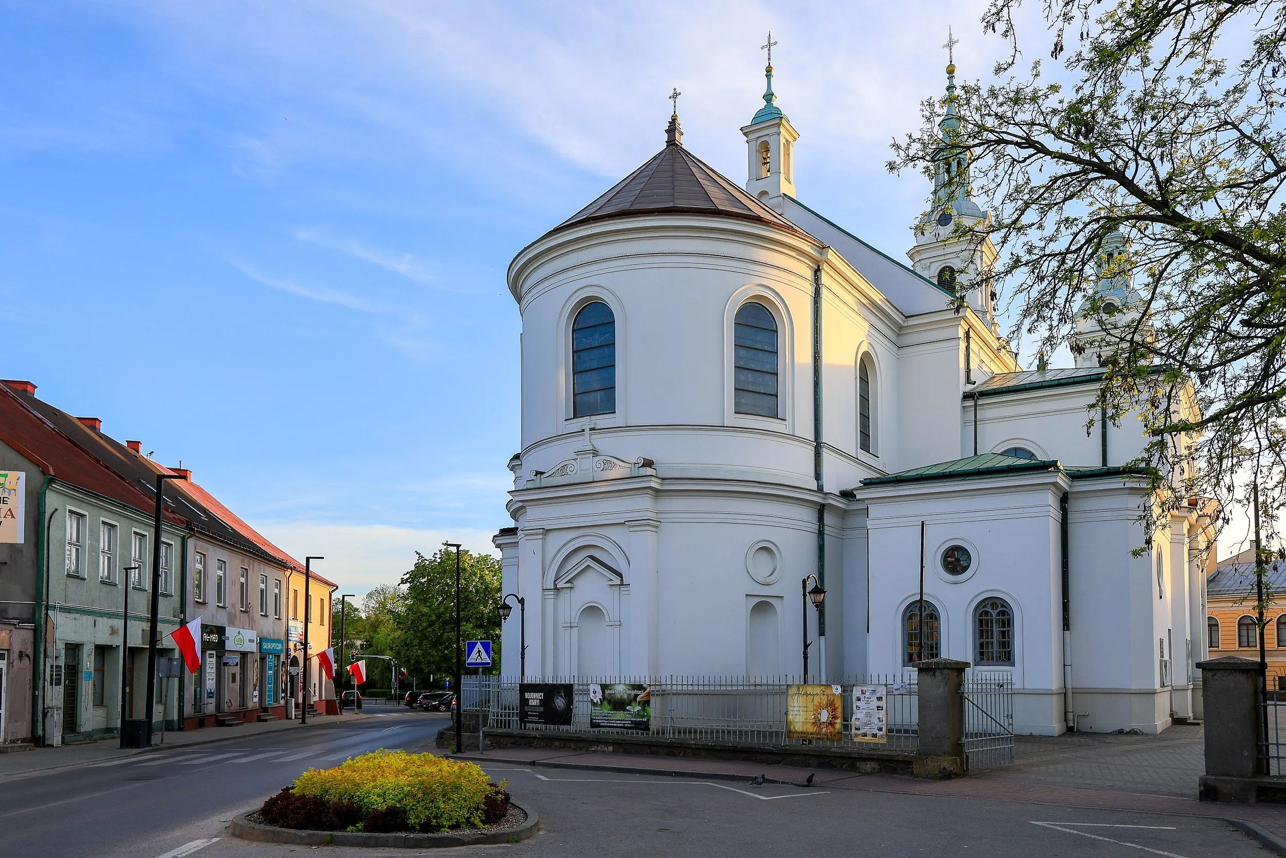 Widok bocznej elewacji Kolegiaty św. Lamberta w Radomsku. Biała świątynia z półkolistą apsydą i wieżami góruje nad zabudową centrum miasta.