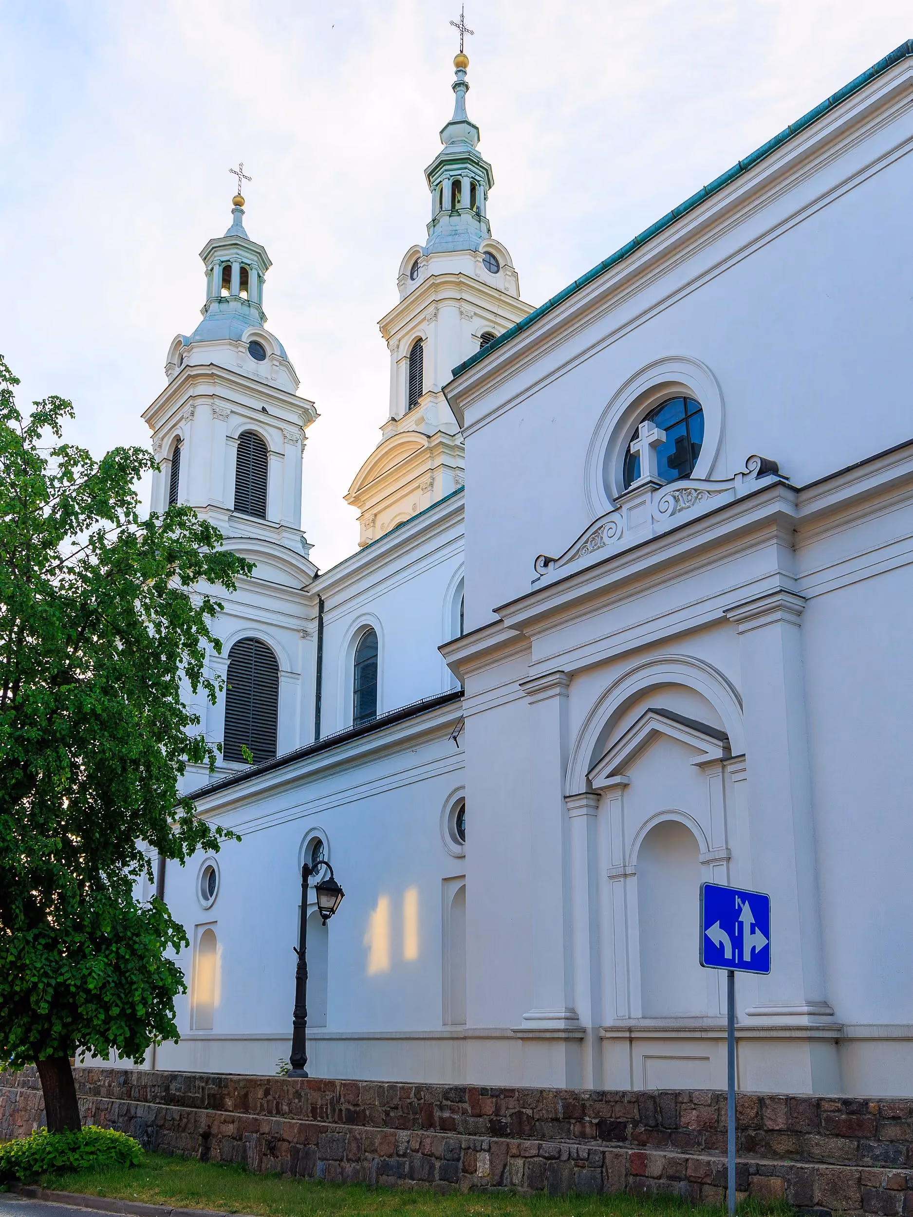 Fragment bocznej elewacji Kolegiaty św. Lamberta w Radomsku. Widoczne białe ściany świątyni, wieże oraz kamienny mur otaczający kościół.