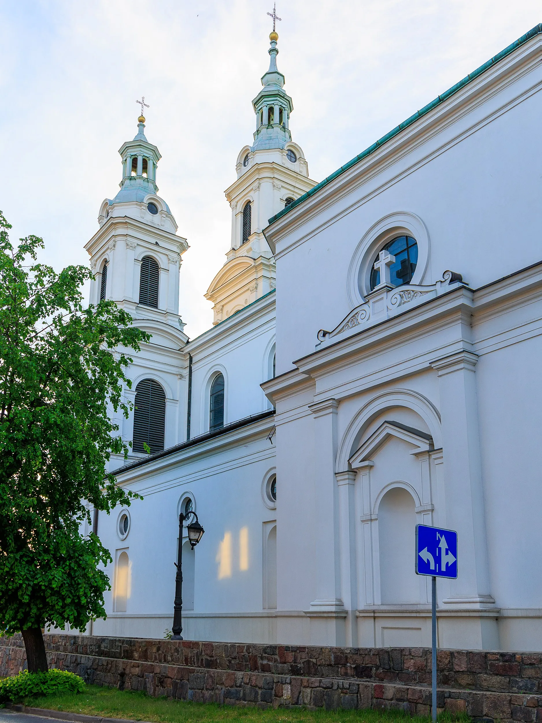 Fragment bocznej elewacji Kolegiaty św. Lamberta w Radomsku. Widoczne białe ściany świątyni, wieże oraz kamienny mur otaczający kościół.