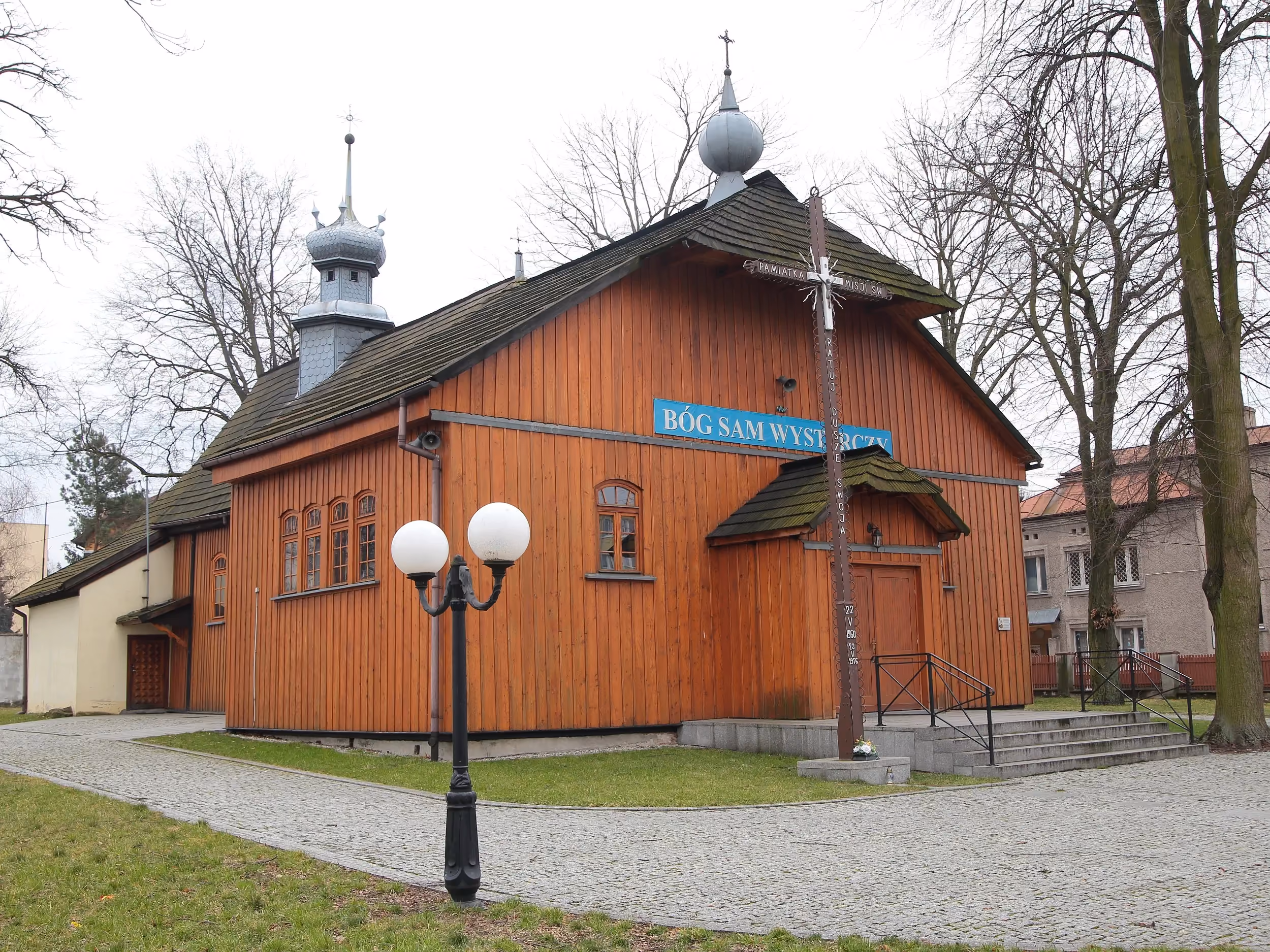 Drewniany kościół św. Marii Magdaleny w Radomsku widziany od frontu. Widoczny gontowy dach, sygnaturka oraz krzyż misyjny przed wejściem.