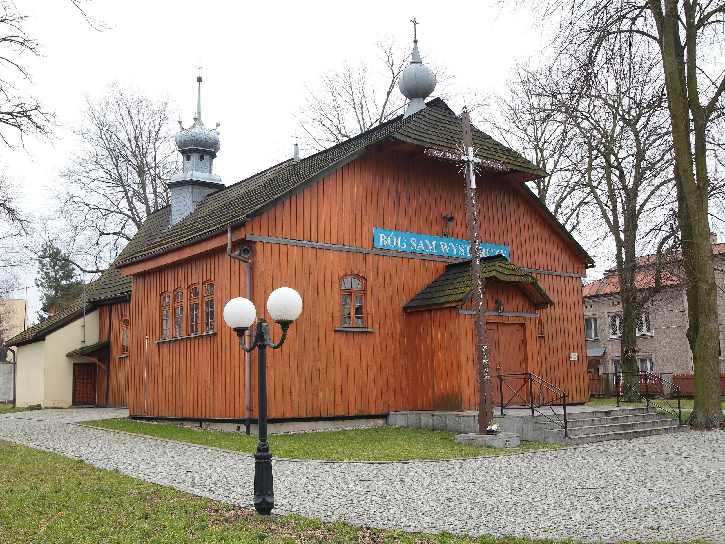 Drewniany kościół św. Marii Magdaleny w Radomsku widziany od frontu. Widoczny gontowy dach, sygnaturka oraz krzyż misyjny przed wejściem.