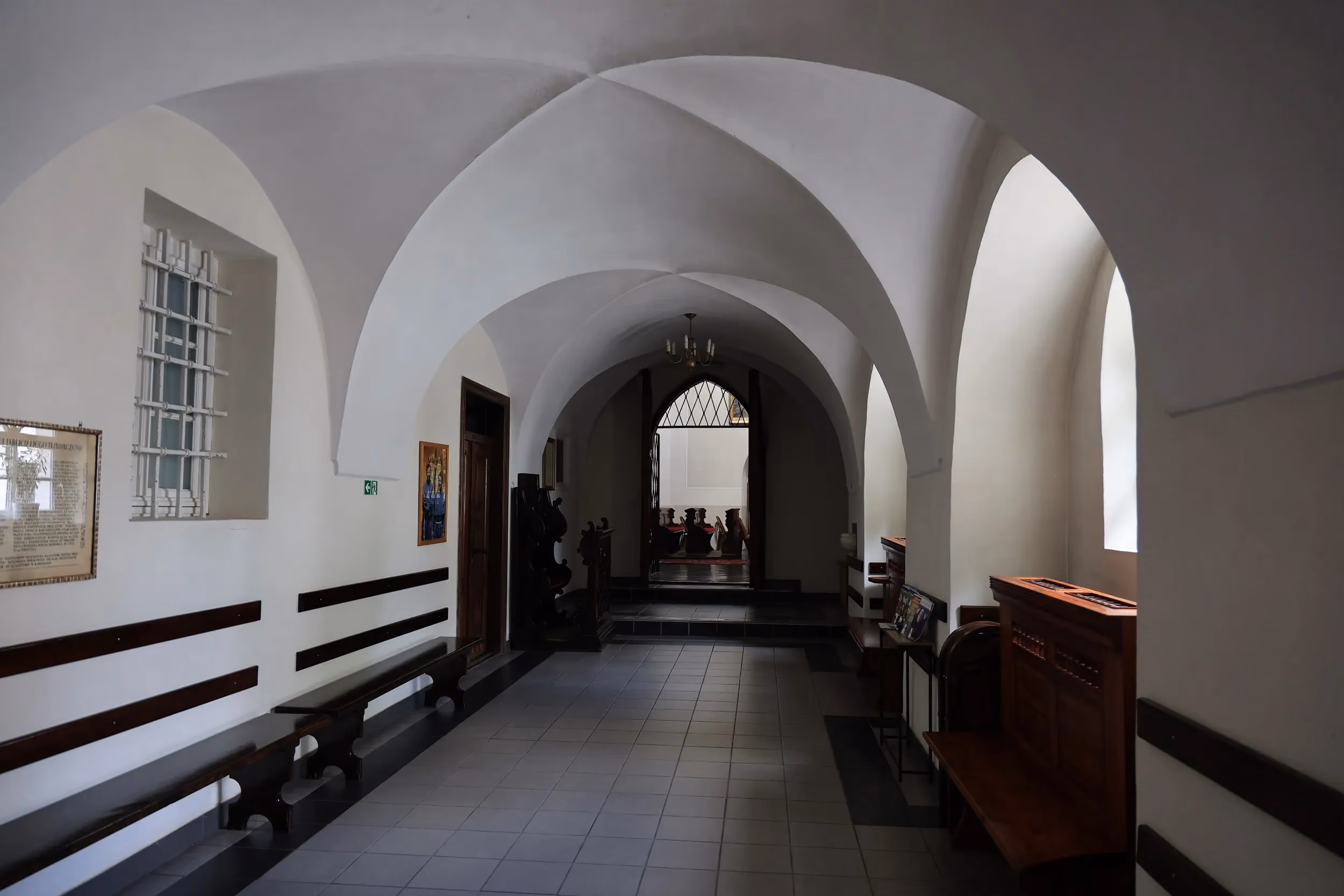 Przestronny krużganek klasztoru franciszkanów w Radomsku z gotyckim sklepieniem, ławkami i wejściem prowadzącym do kościoła.