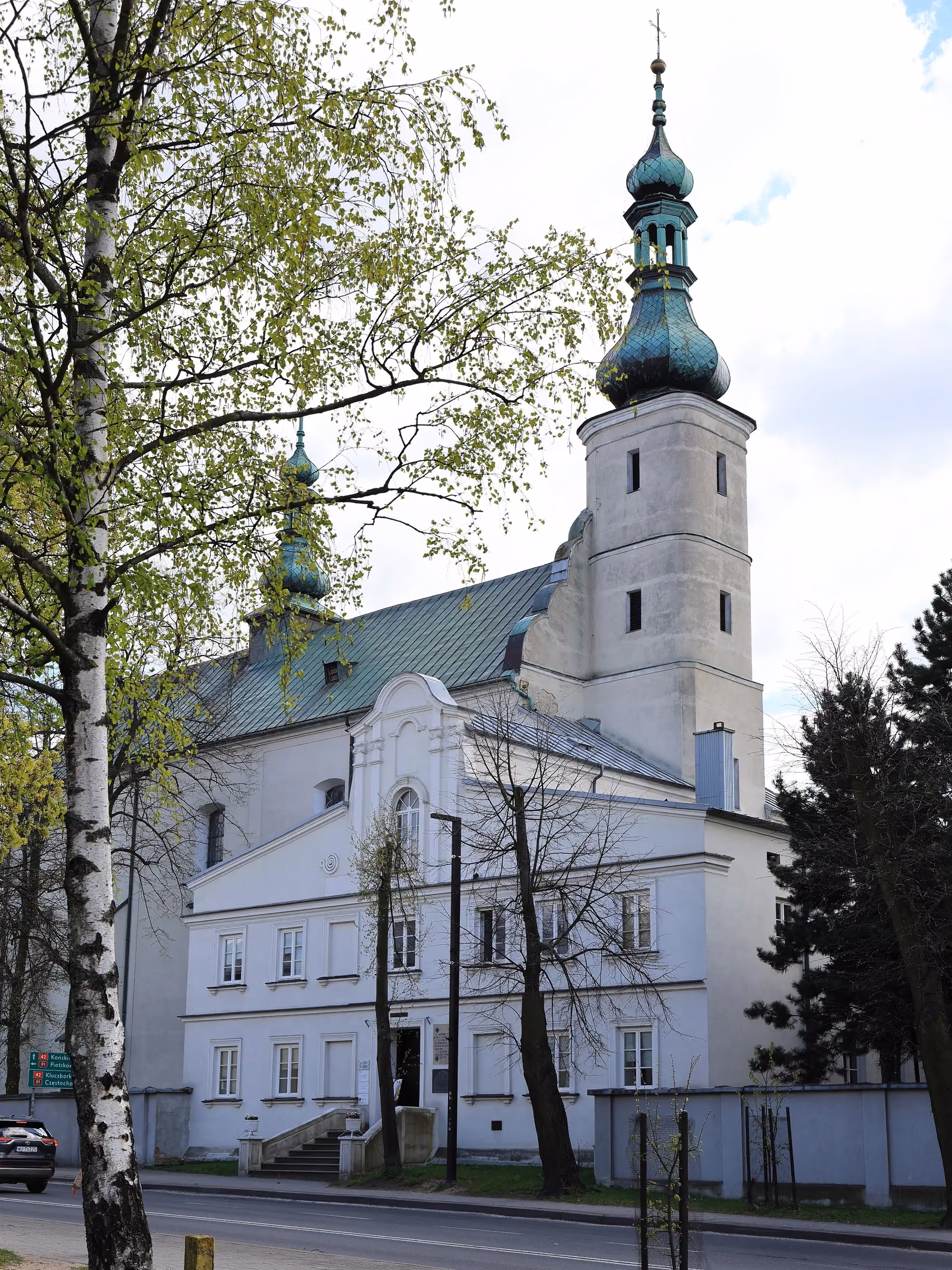 Widok klasztoru franciszkanów w Radomsku na tle kościoła św. Lamberta. Widoczne barokowe wieże świątyni oraz drzewa otaczające kompleks.