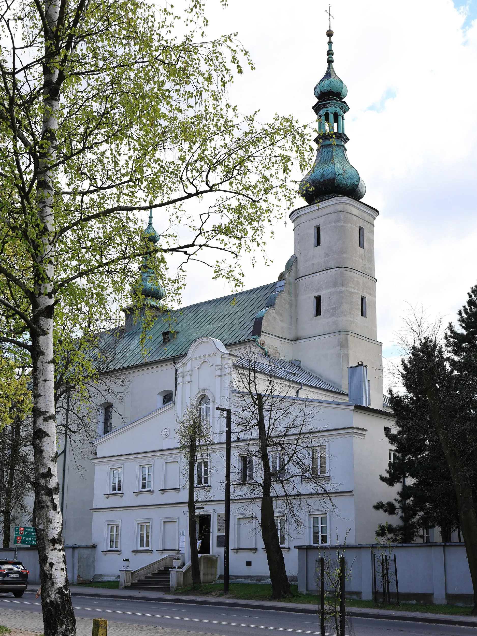 Widok klasztoru franciszkanów w Radomsku na tle kościoła św. Lamberta. Widoczne barokowe wieże świątyni oraz drzewa otaczające kompleks.