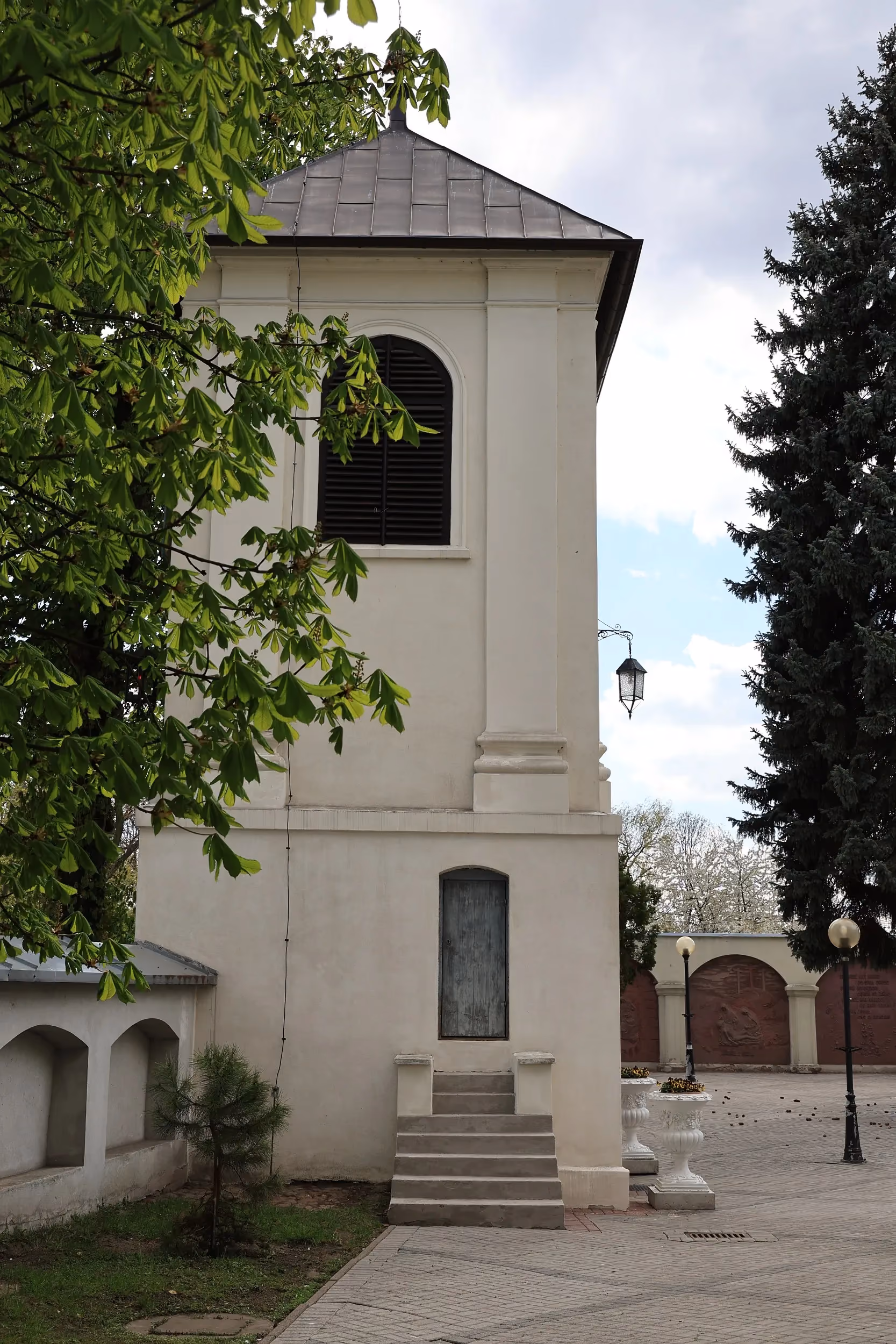 Boczny widok dzwonnicy klasztornej w Radomsku z wejściem, schodami oraz placem otoczonym drzewami.