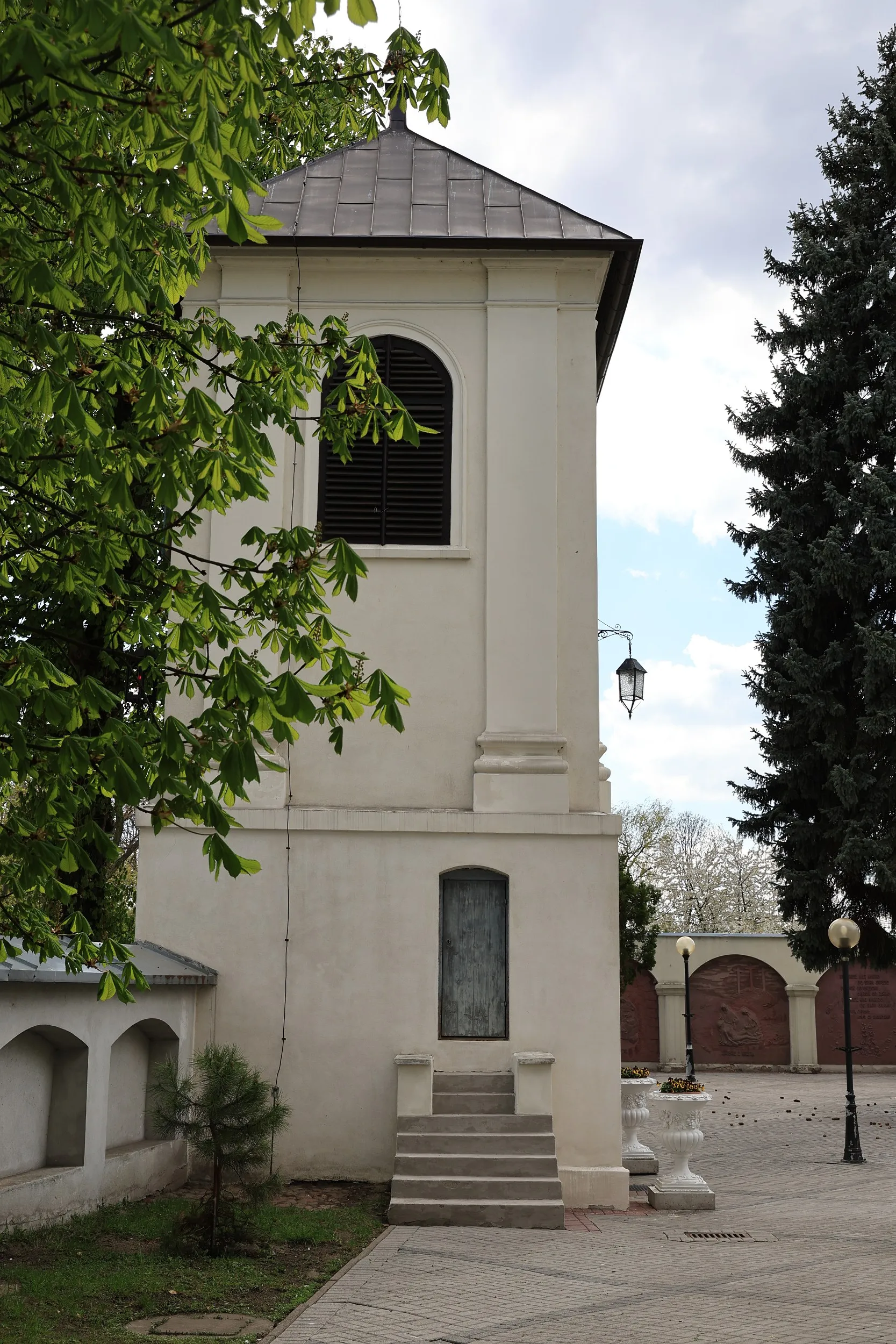 Boczny widok dzwonnicy klasztornej w Radomsku z wejściem, schodami oraz placem otoczonym drzewami.
