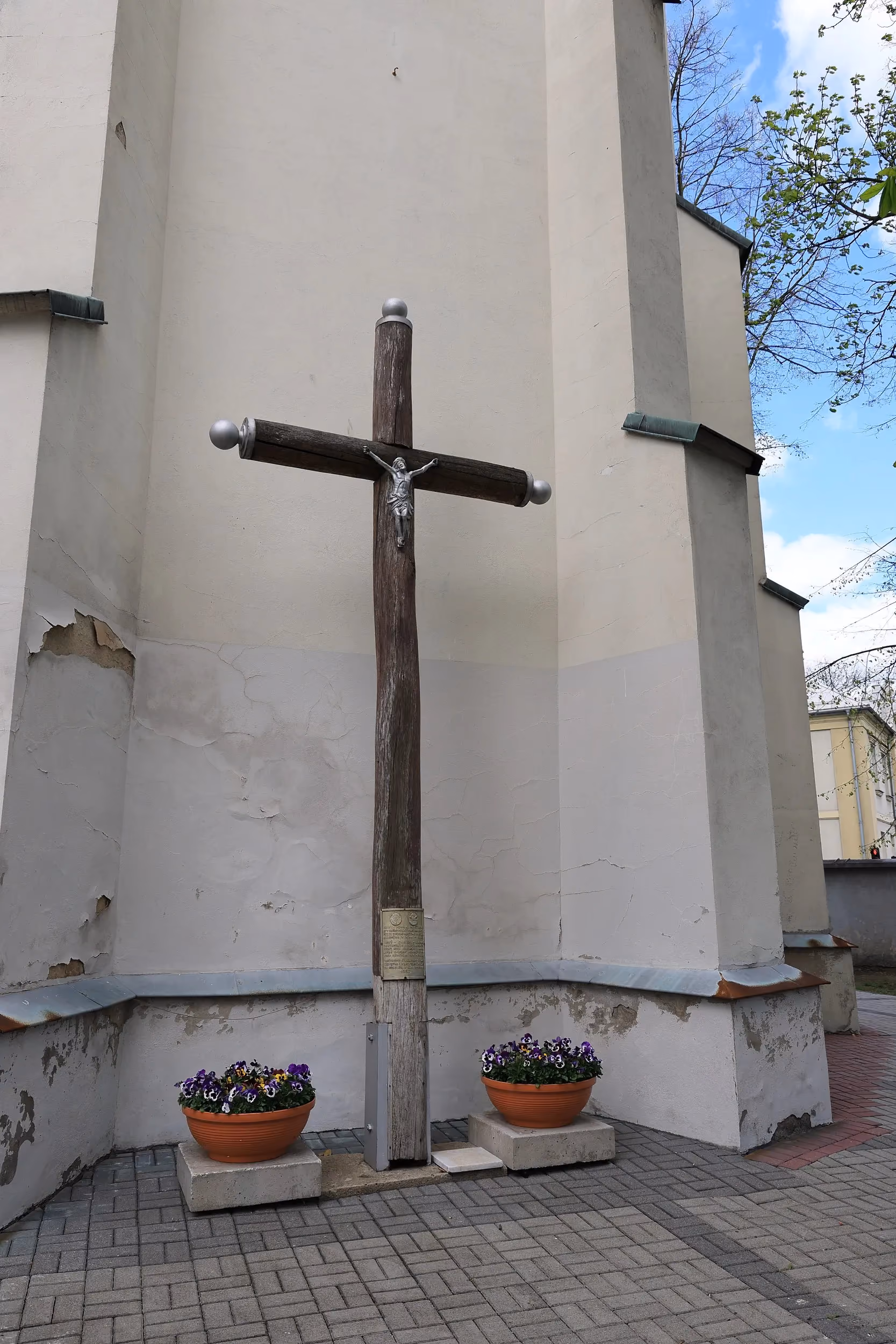 Drewniany krzyż powstańczy z figurą Chrystusa ustawiony przy murze klasztoru franciszkanów w Radomsku, otoczony kwiatami w donicach.