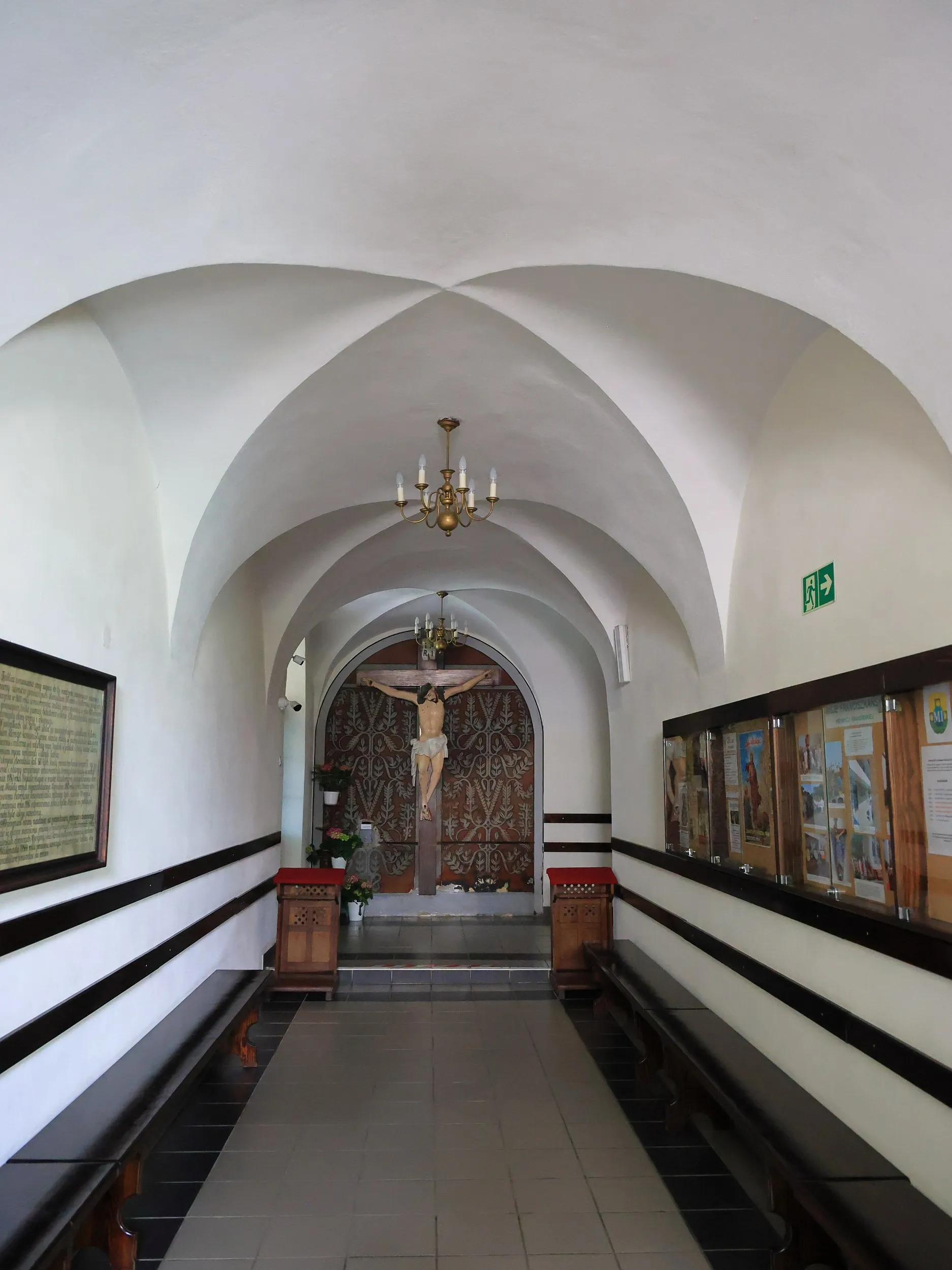 Korytarz klasztorny z kolebkowym sklepieniem, ławkami oraz krucyfiksem umieszczonym na końcu przejścia.