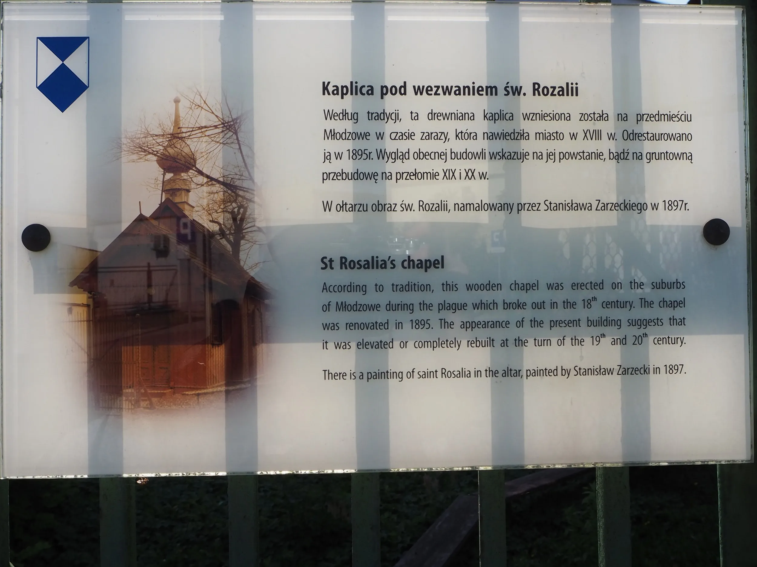 Tablica informacyjna przy kaplicy św. Rozalii w Radomsku z opisem historii drewnianej świątyni oraz fotografią obiektu.