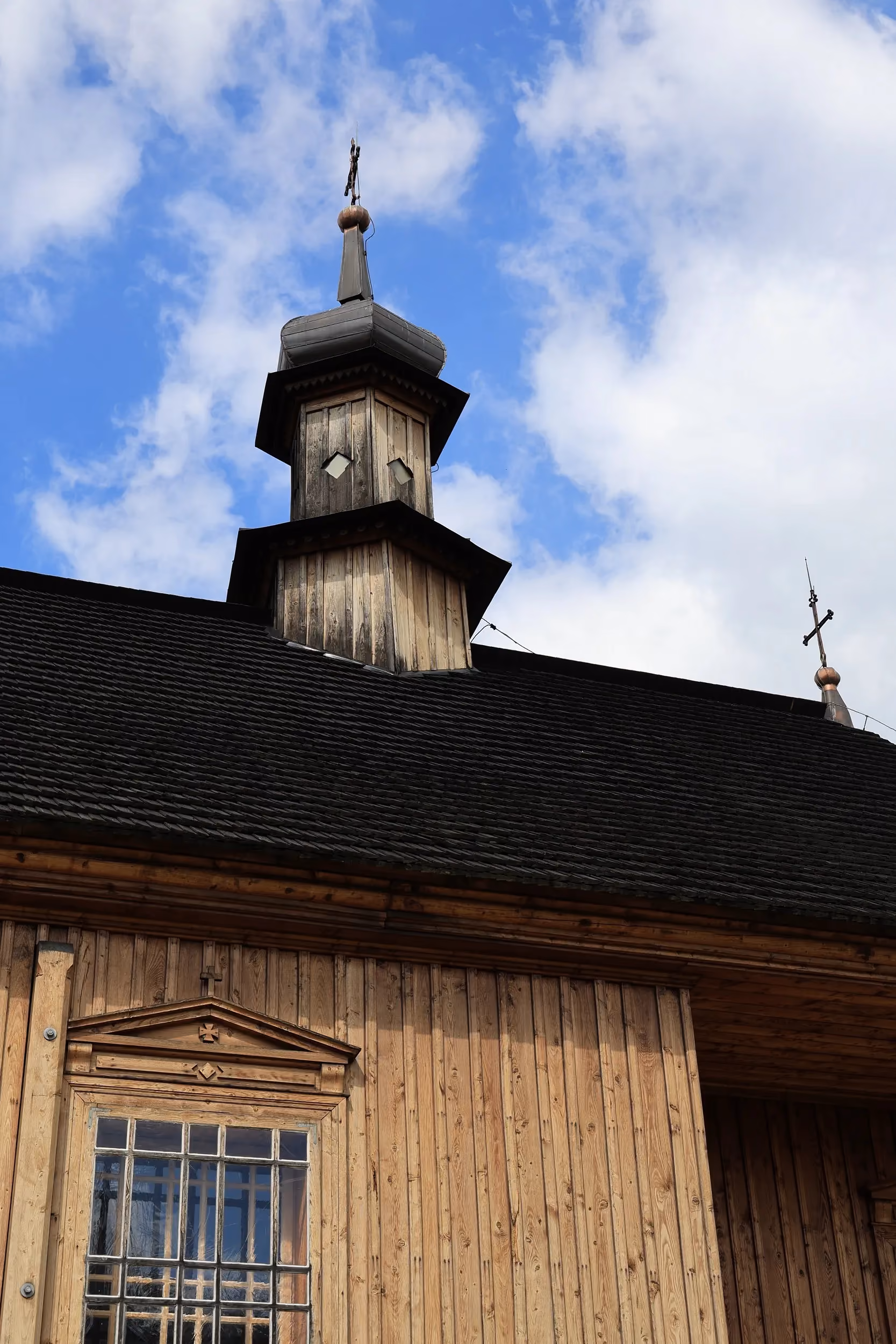Drewniana sygnaturka z cebulastym hełmem i krzyżem wieńcząca dach kościoła św. Rocha. W tle widoczne niebo z chmurami.