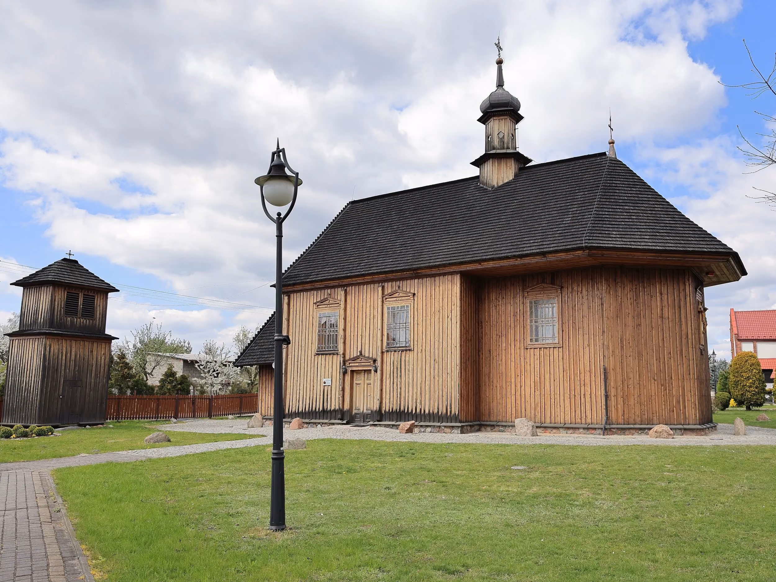 Kościół św. Rocha w Radomsku wraz z wolnostojącą drewnianą dzwonnicą. Obiekty otacza zadbany teren z alejkami i zielenią.