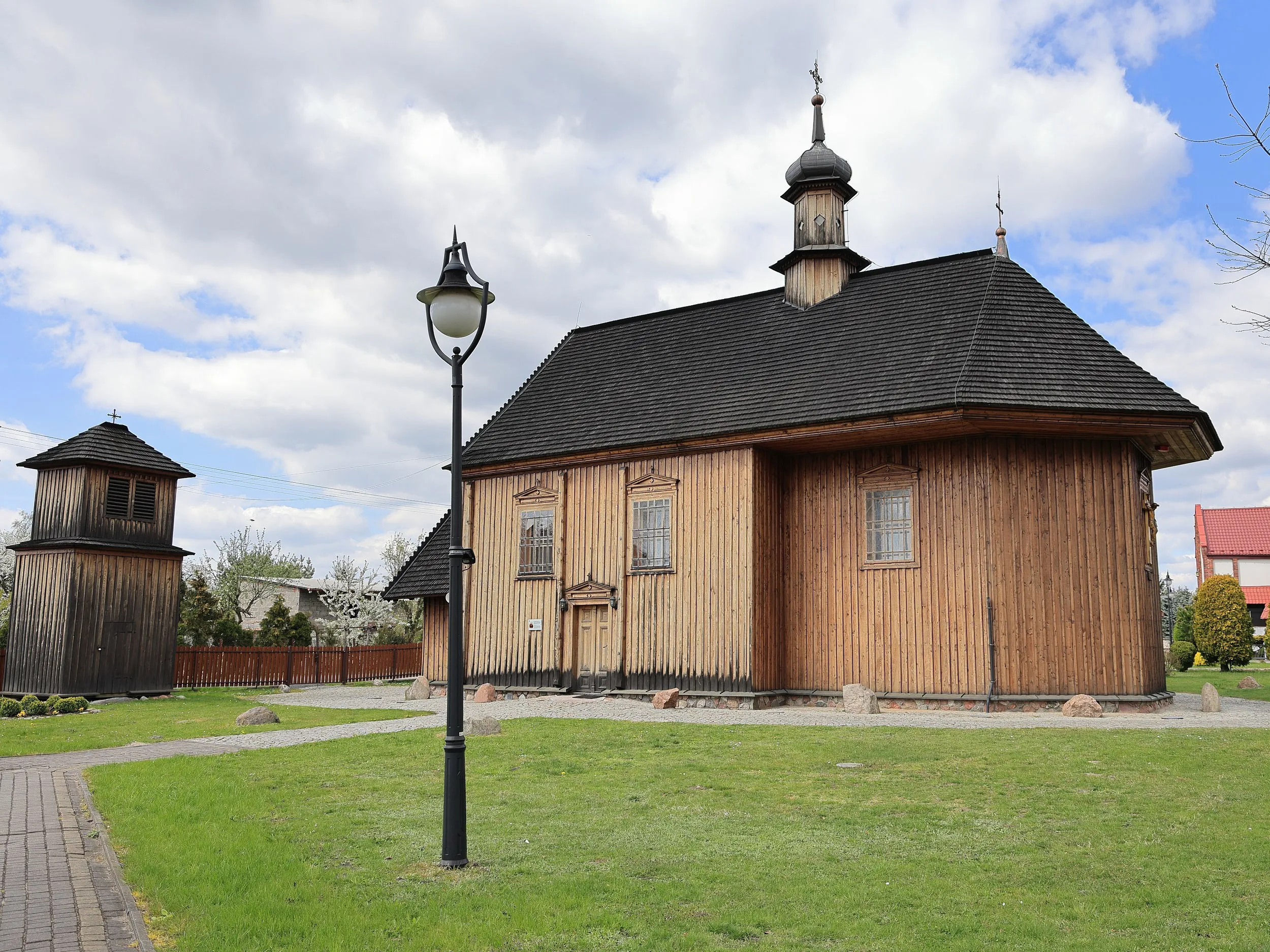 Kościół św. Rocha w Radomsku wraz z wolnostojącą drewnianą dzwonnicą. Obiekty otacza zadbany teren z alejkami i zielenią.