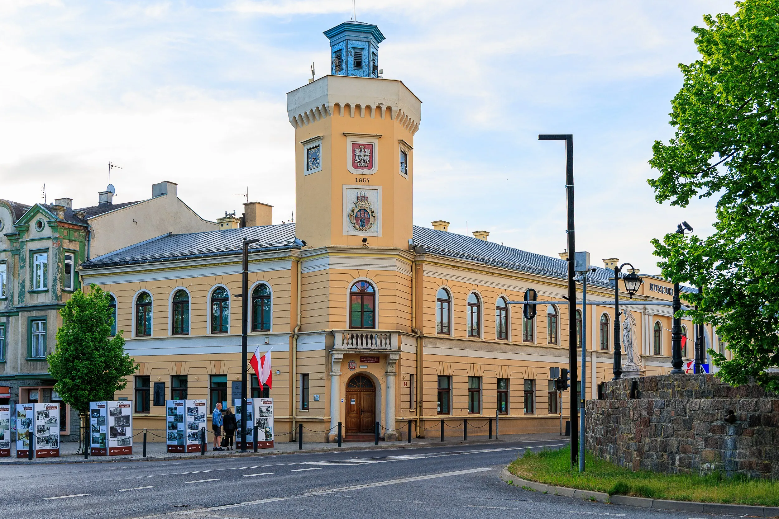 Front zabytkowego ratusza w Radomsku z centralnie usytuowaną wieżą. Przed budynkiem znajdują się flagi oraz plansze wystawowe.
