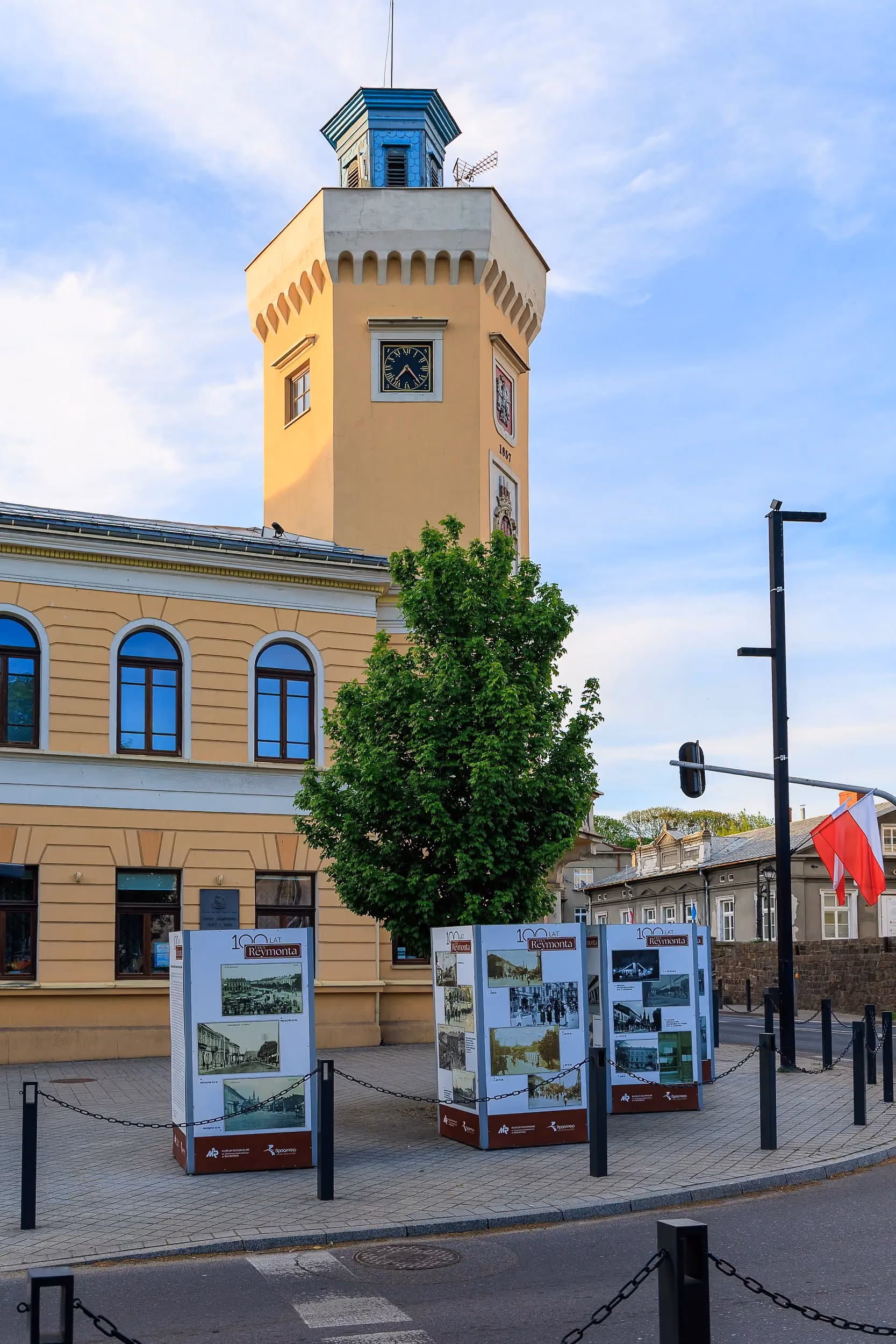 Wieża zabytkowego ratusza w Radomsku z zegarem i herbami. Na pierwszym planie widoczne są plansze prezentujące historię miasta.