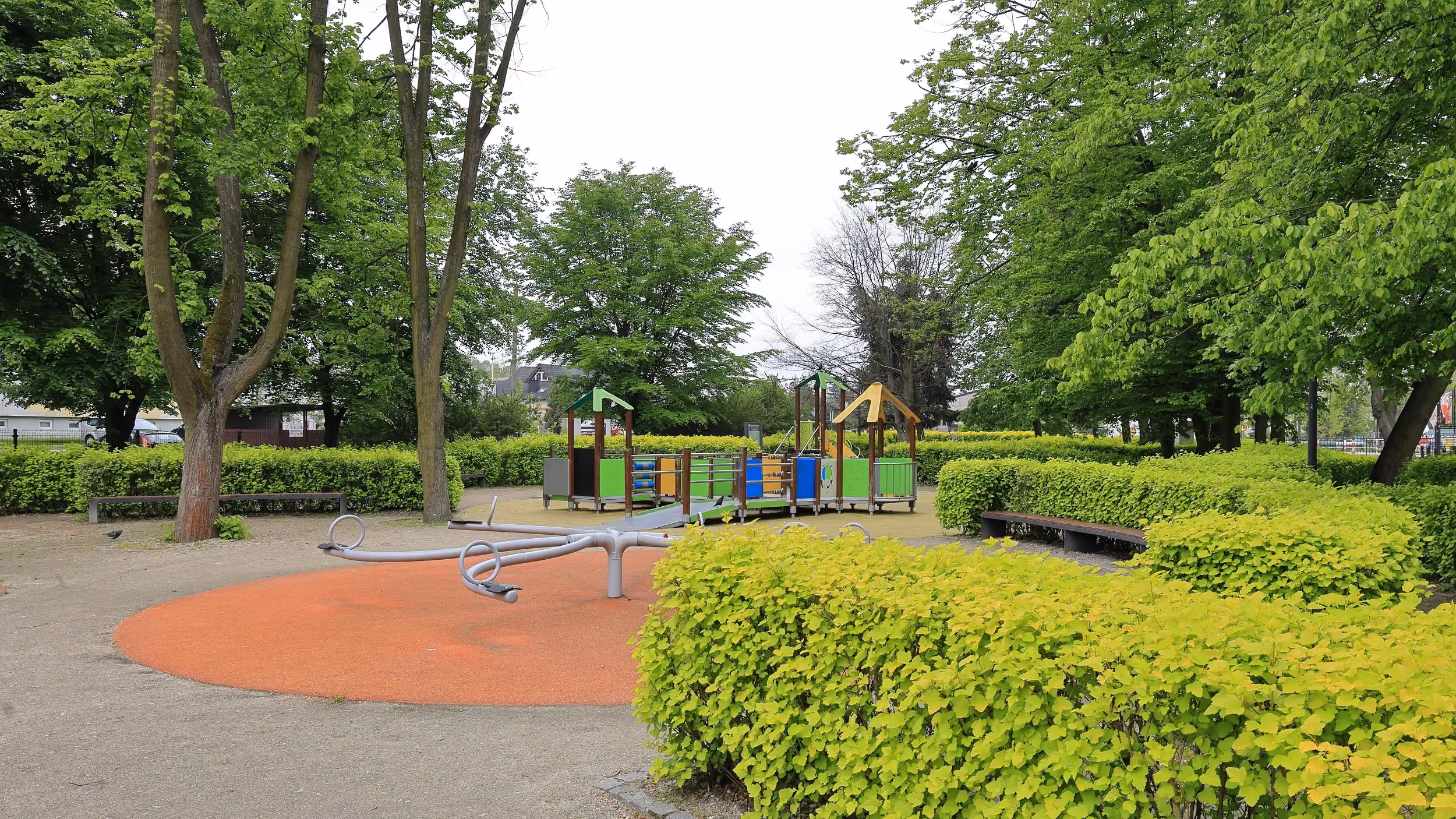 Plac zabaw w zabytkowym parku w Radomsku. Wśród zieleni znajdują się urządzenia rekreacyjne dla dzieci oraz miejsca do odpoczynku.