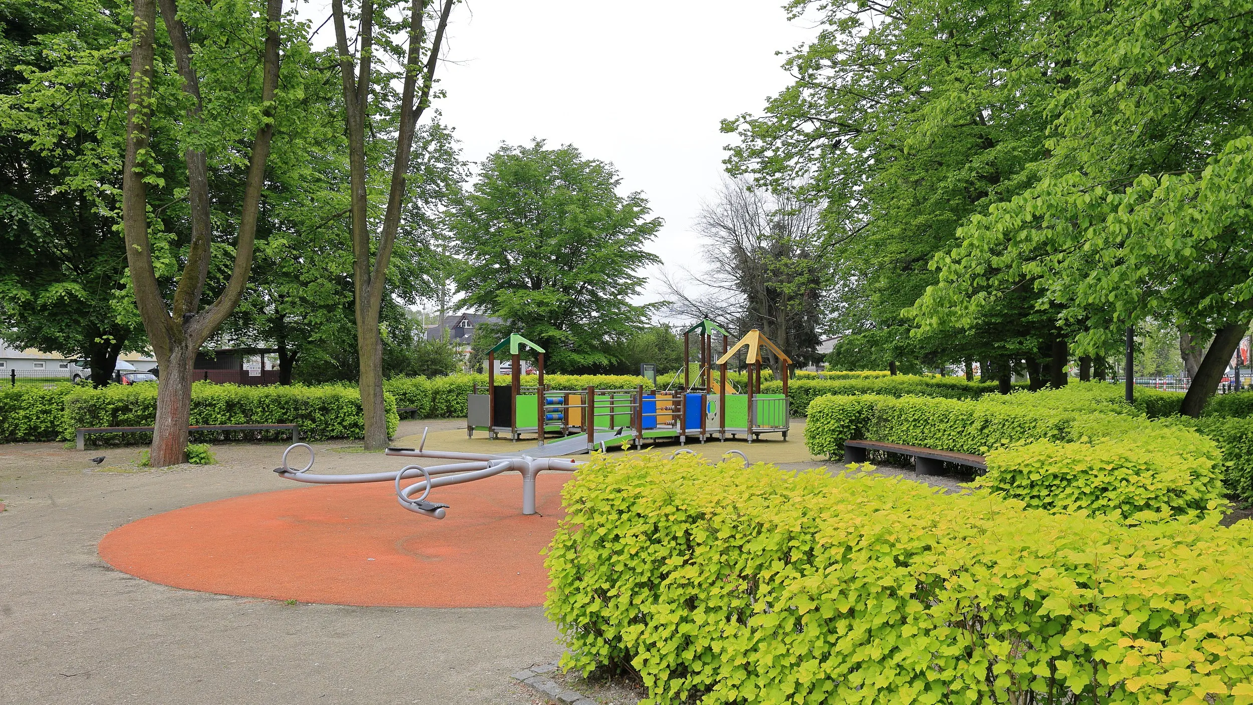 Plac zabaw w zabytkowym parku w Radomsku. Wśród zieleni znajdują się urządzenia rekreacyjne dla dzieci oraz miejsca do odpoczynku.