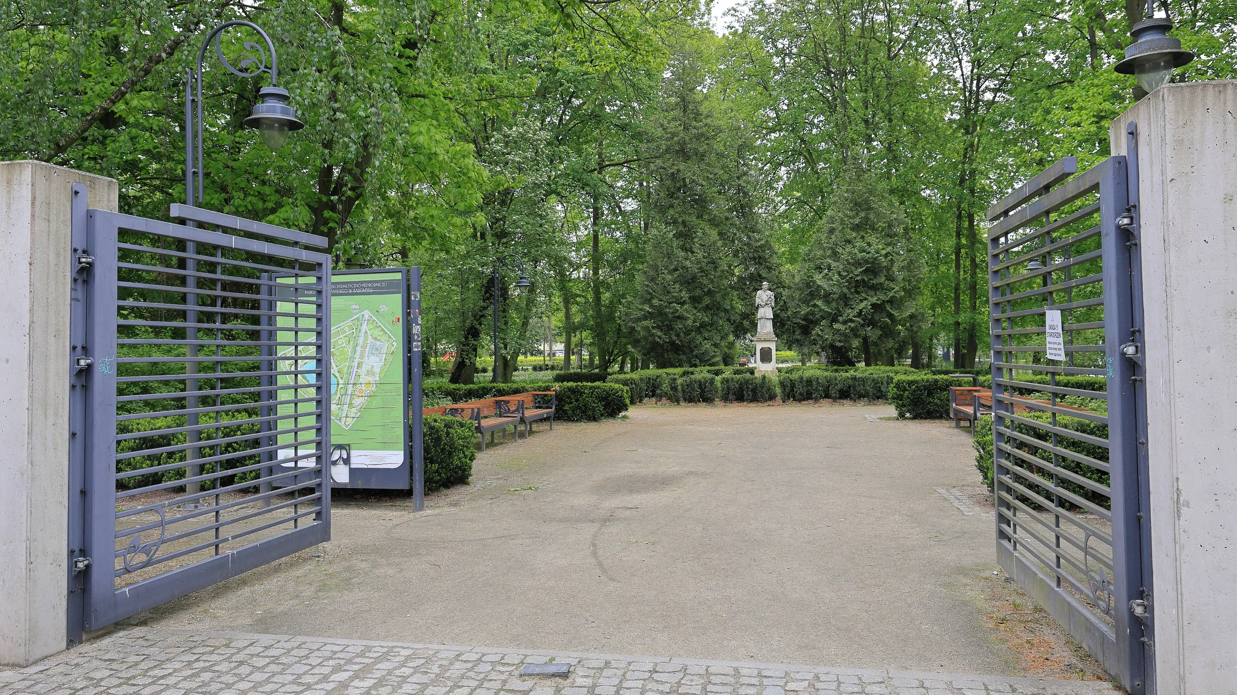 Główne wejście do zabytkowego parku w Radomsku. Otwarta brama prowadzi do alejek spacerowych, otoczonych drzewami i zadbaną zielenią.
