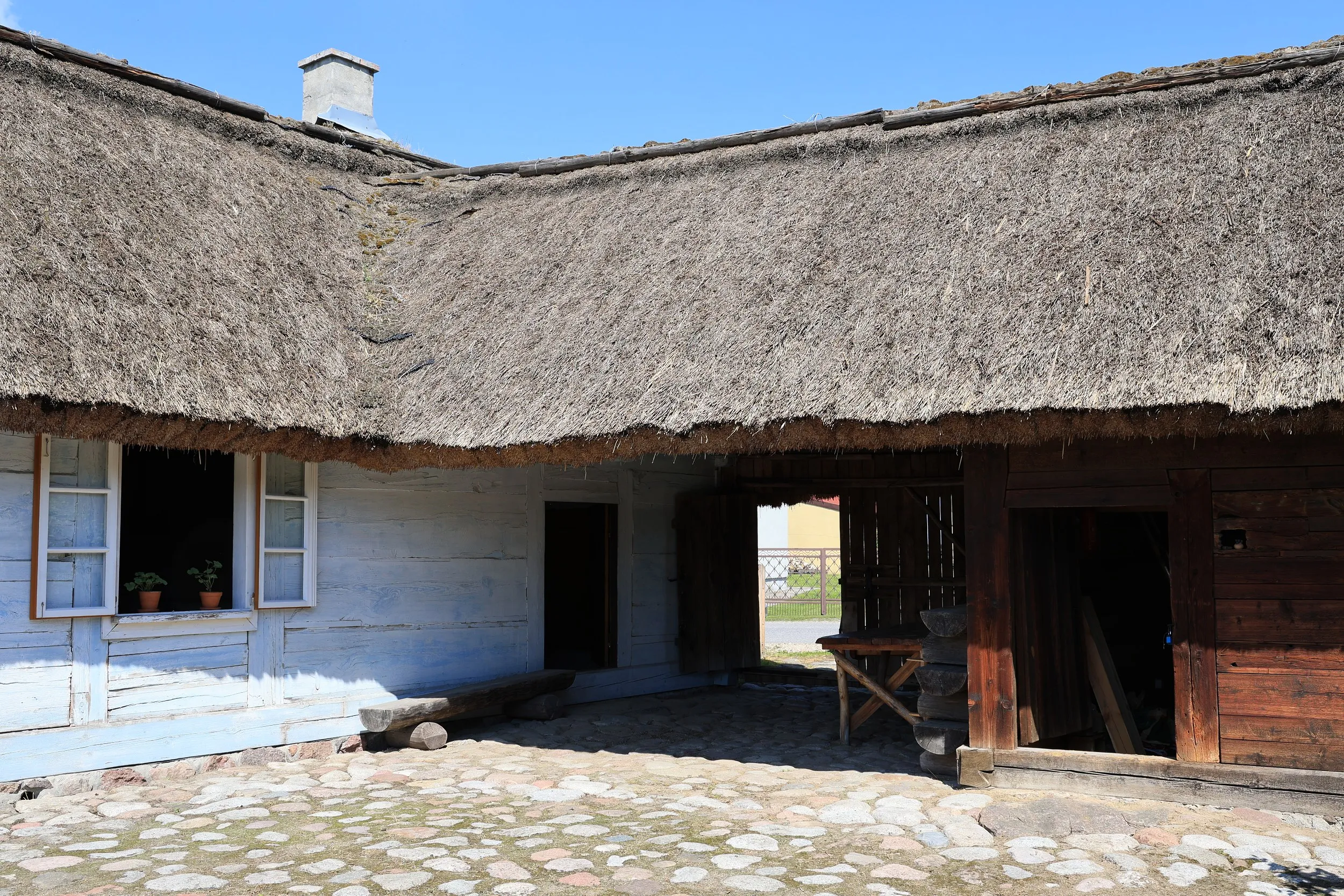 Dziedziniec zabytkowej chaty tatarskiej w Radomsku z brukowaną nawierzchnią, drewnianą zabudową i strzechą.