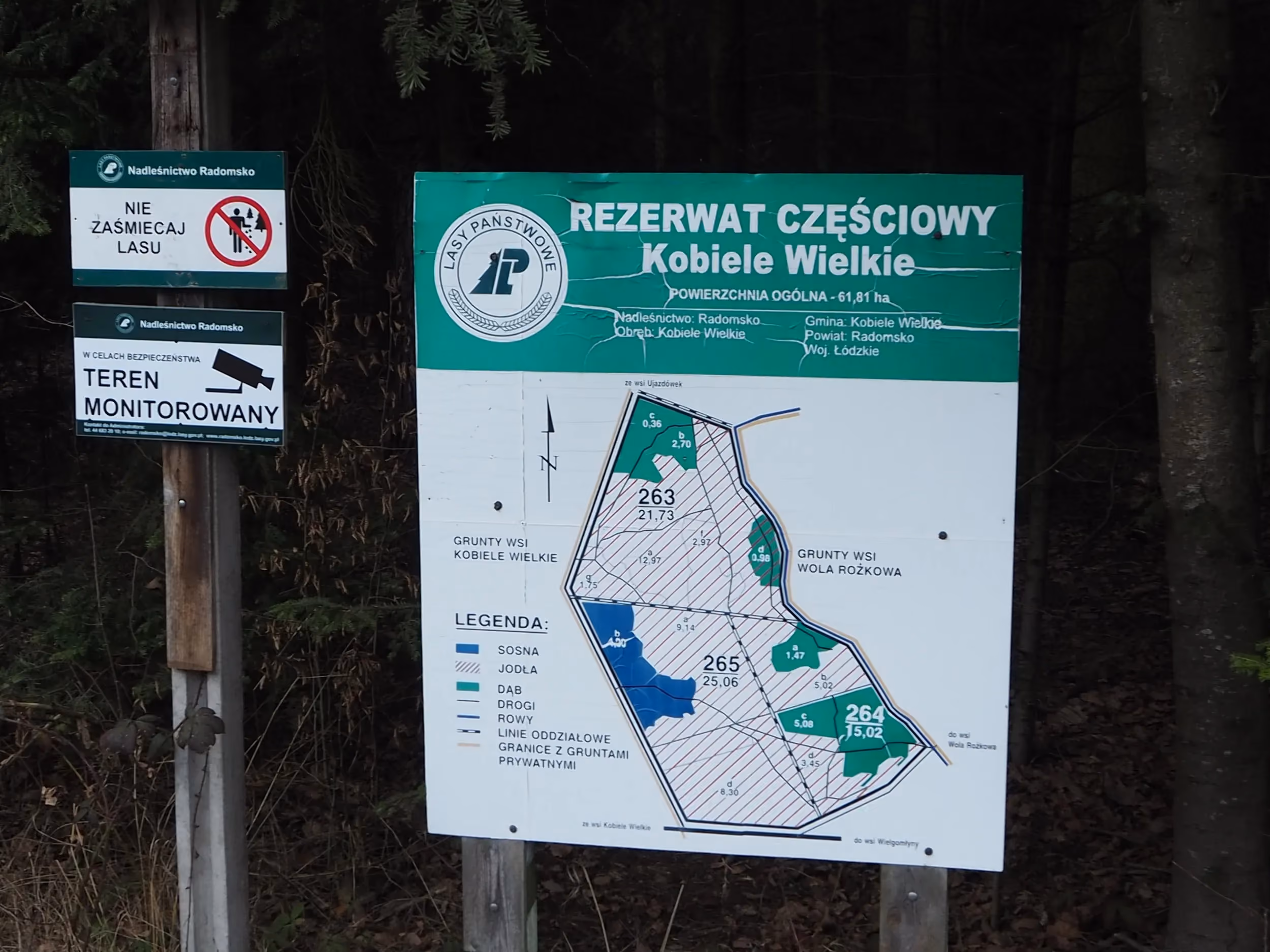 Tablica informacyjna rezerwatu częściowego „Kobiele Wielkie” z mapą obszaru i zasadami ochrony, ustawiona przy skraju lasu.