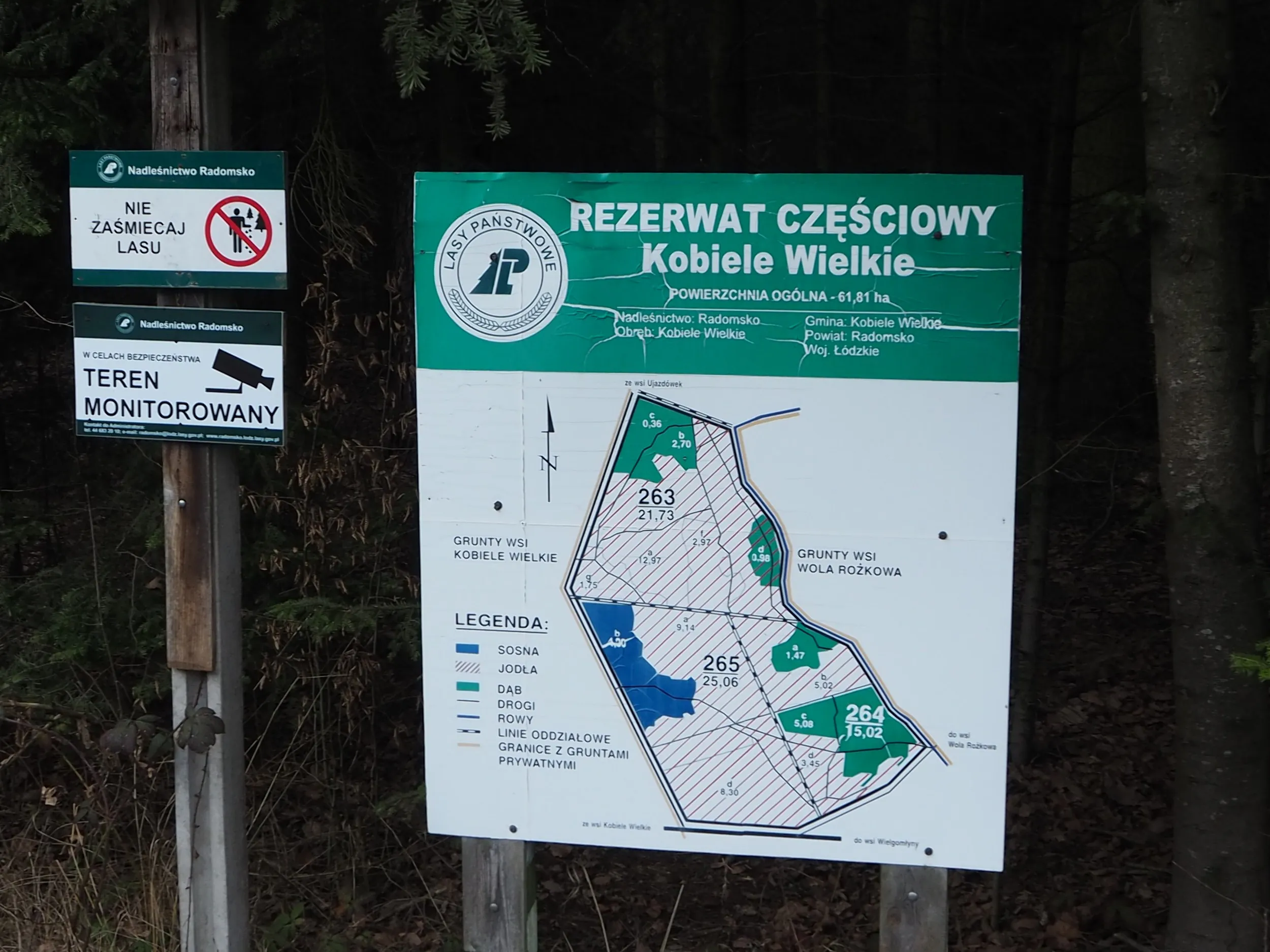 Tablica informacyjna rezerwatu częściowego „Kobiele Wielkie” z mapą obszaru i zasadami ochrony, ustawiona przy skraju lasu.