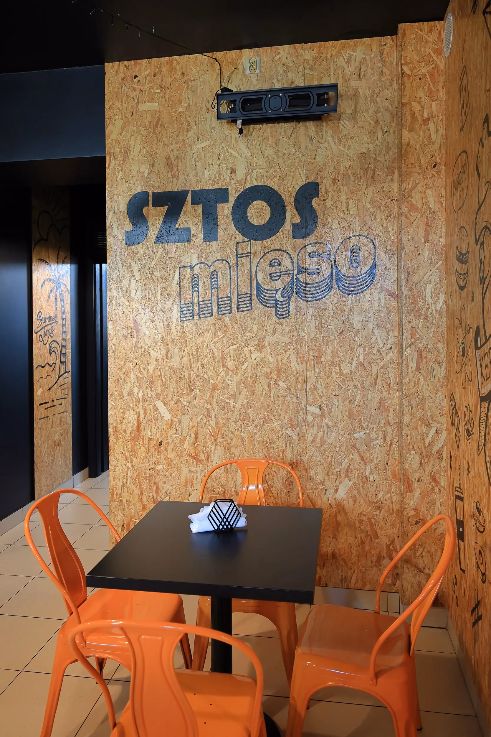 Strefa restauracyjna Sztos Mięso w Przedborzu z industrialnym wystrojem, czarnym stolikiem, pomarańczowymi krzesłami i drewnianą ścianą z logo lokalu.