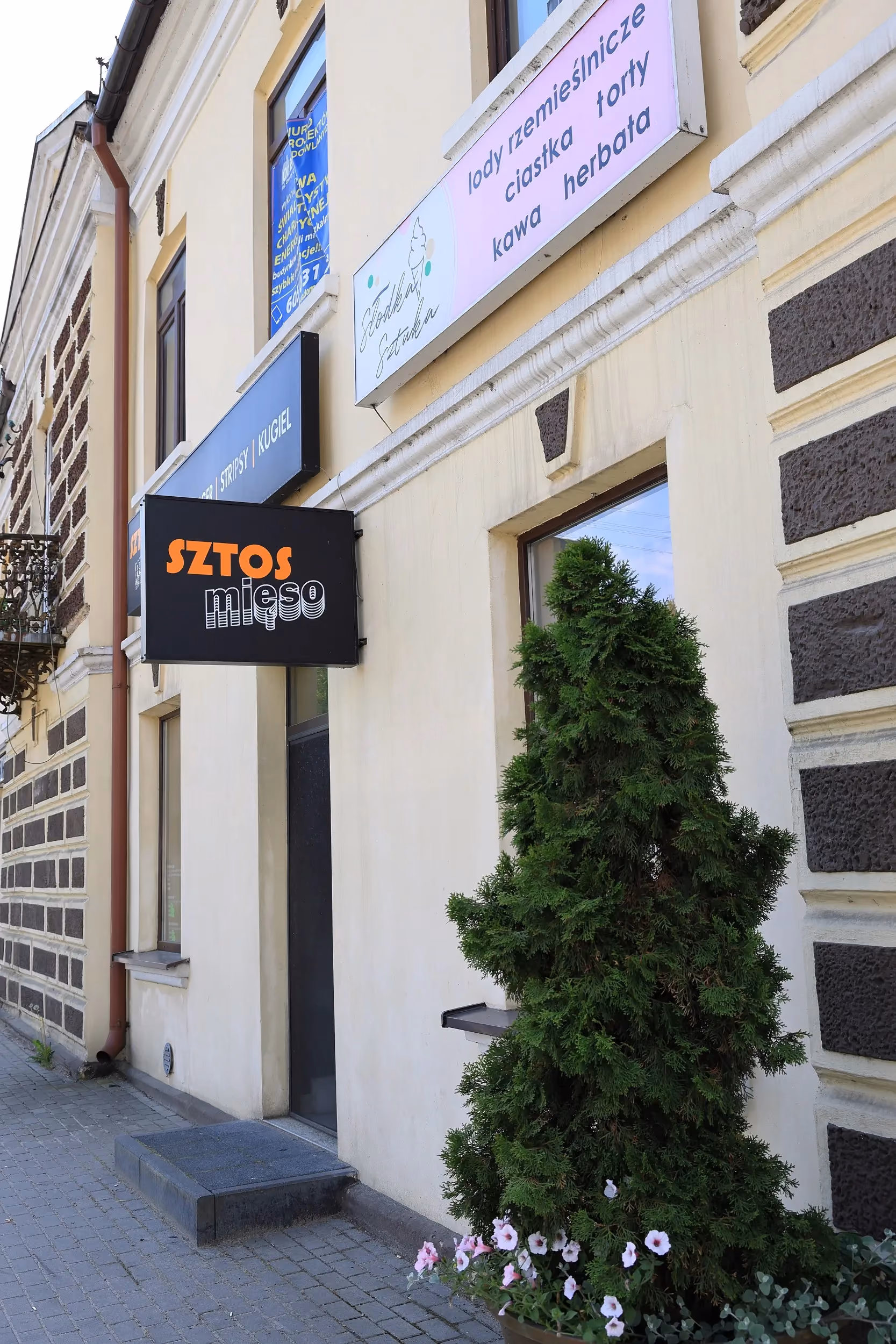 Wejście do restauracji Sztos Mięso w centrum Przedborza z widocznym szyldem lokalu i dekoracyjną zielenią.